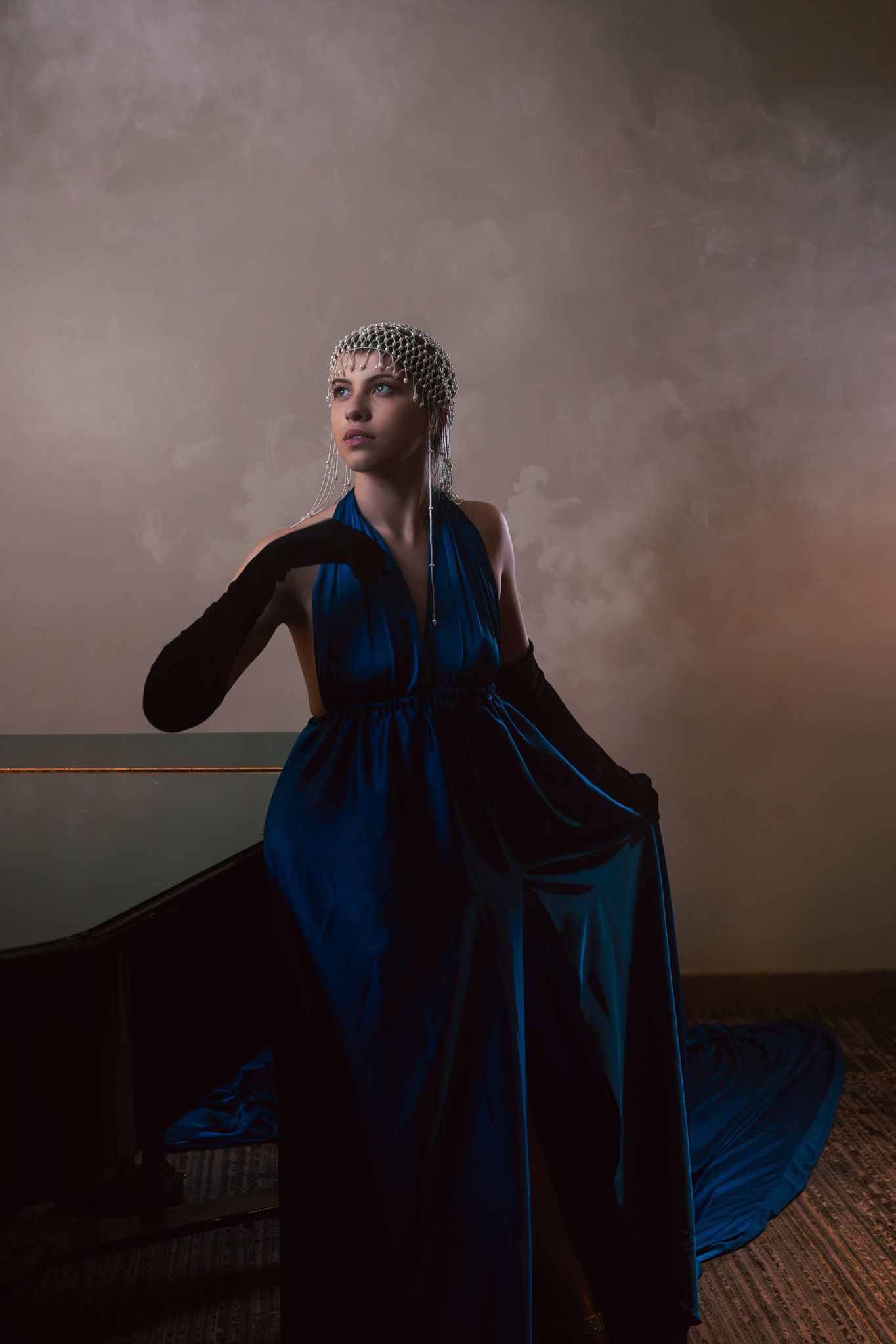 Elegant woman in a blue velvet gown and pearl headpiece poses dramatically in a smoky, dimly lit setting.