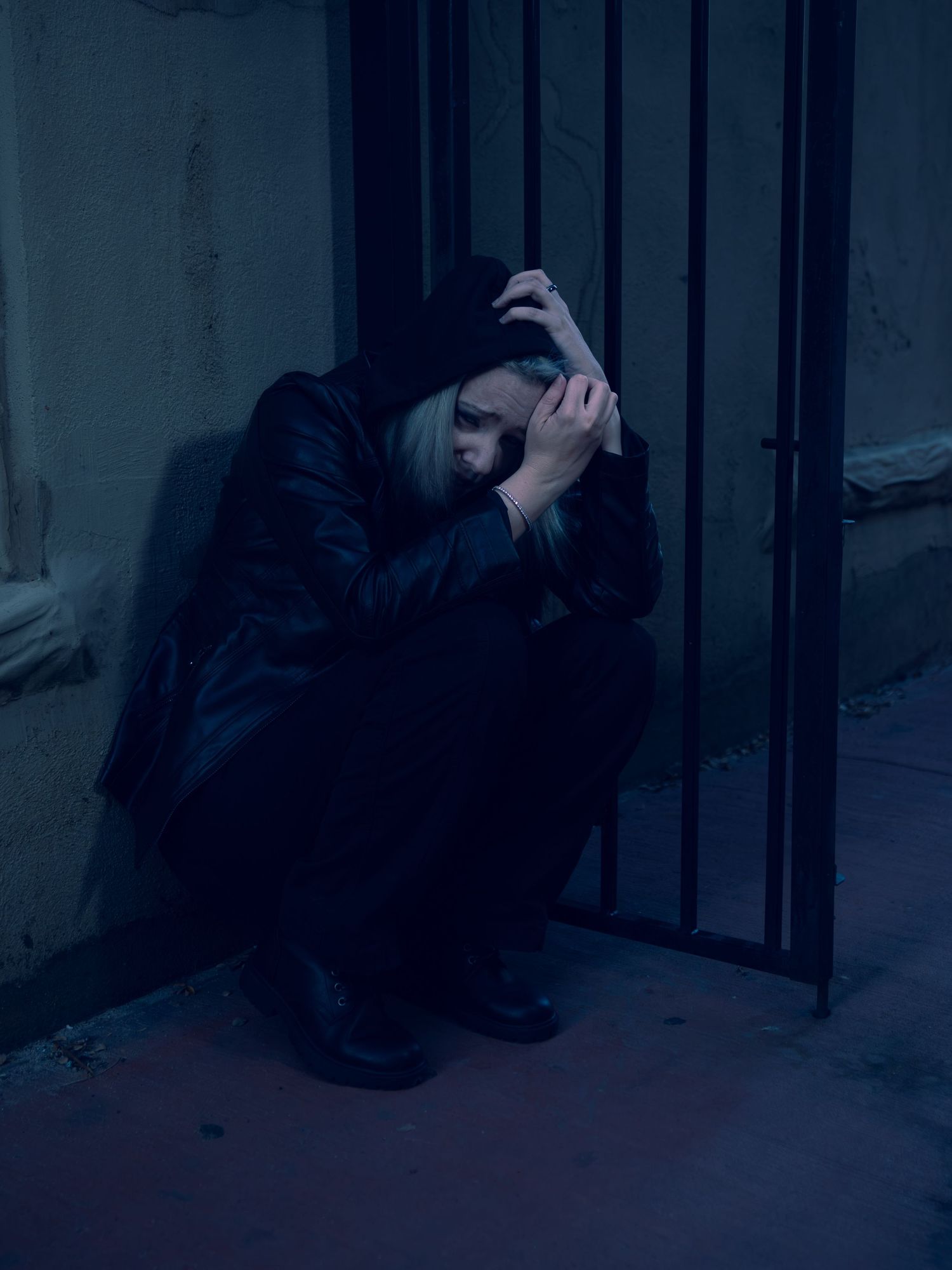 Person in dark clothing sitting alone against metal bars in dim blue light, appearing distressed.