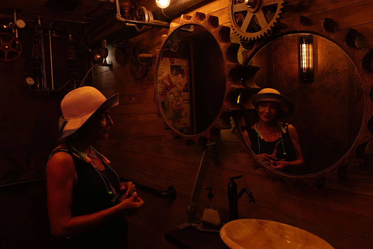 Woman in yellow hat examines reflection in round mirror inside dimly lit steampunk-themed wooden room with gears.