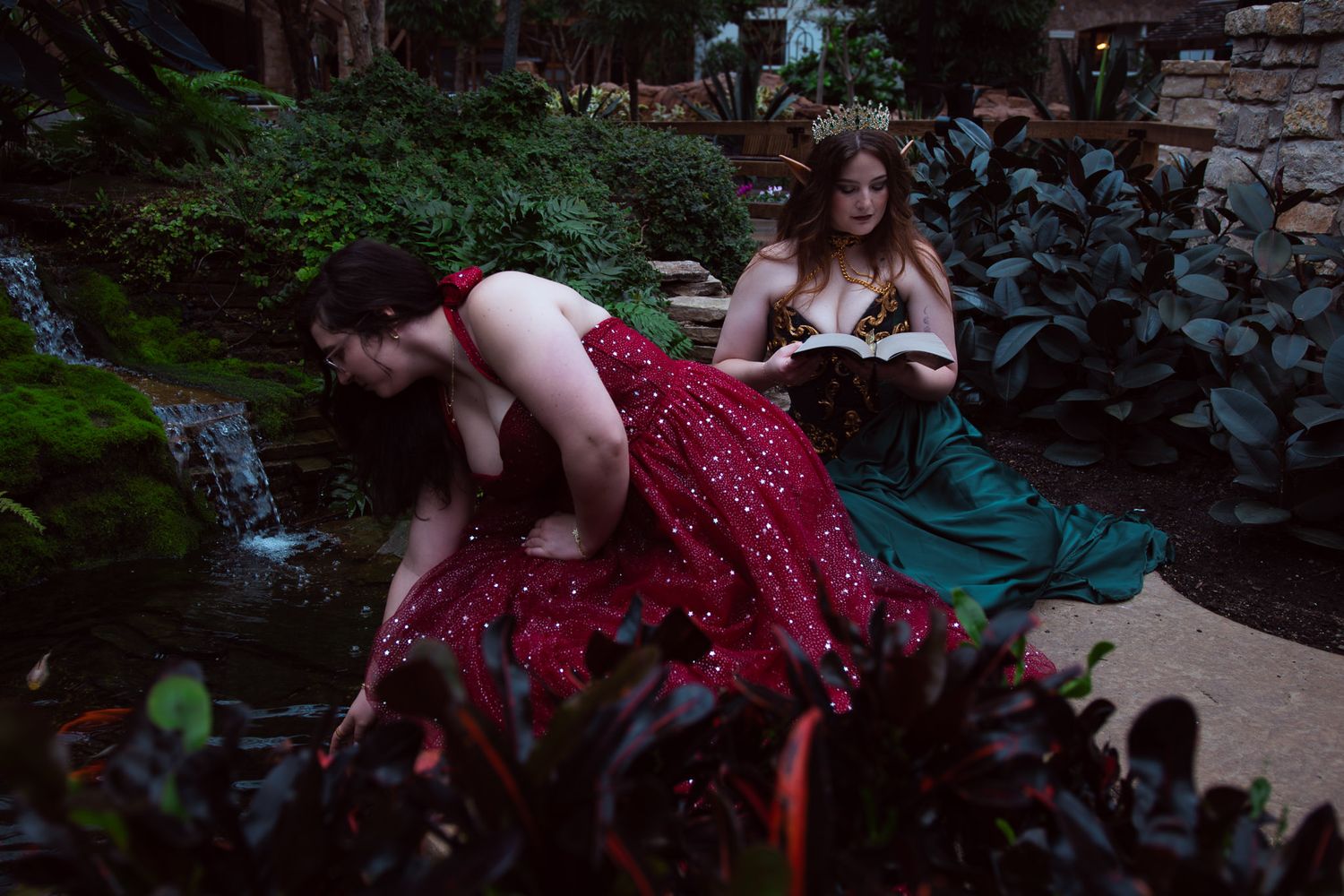 Two women in elegant red and green gowns pose near a garden waterfall surrounded by lush greenery at dusk.  One is reading a book.