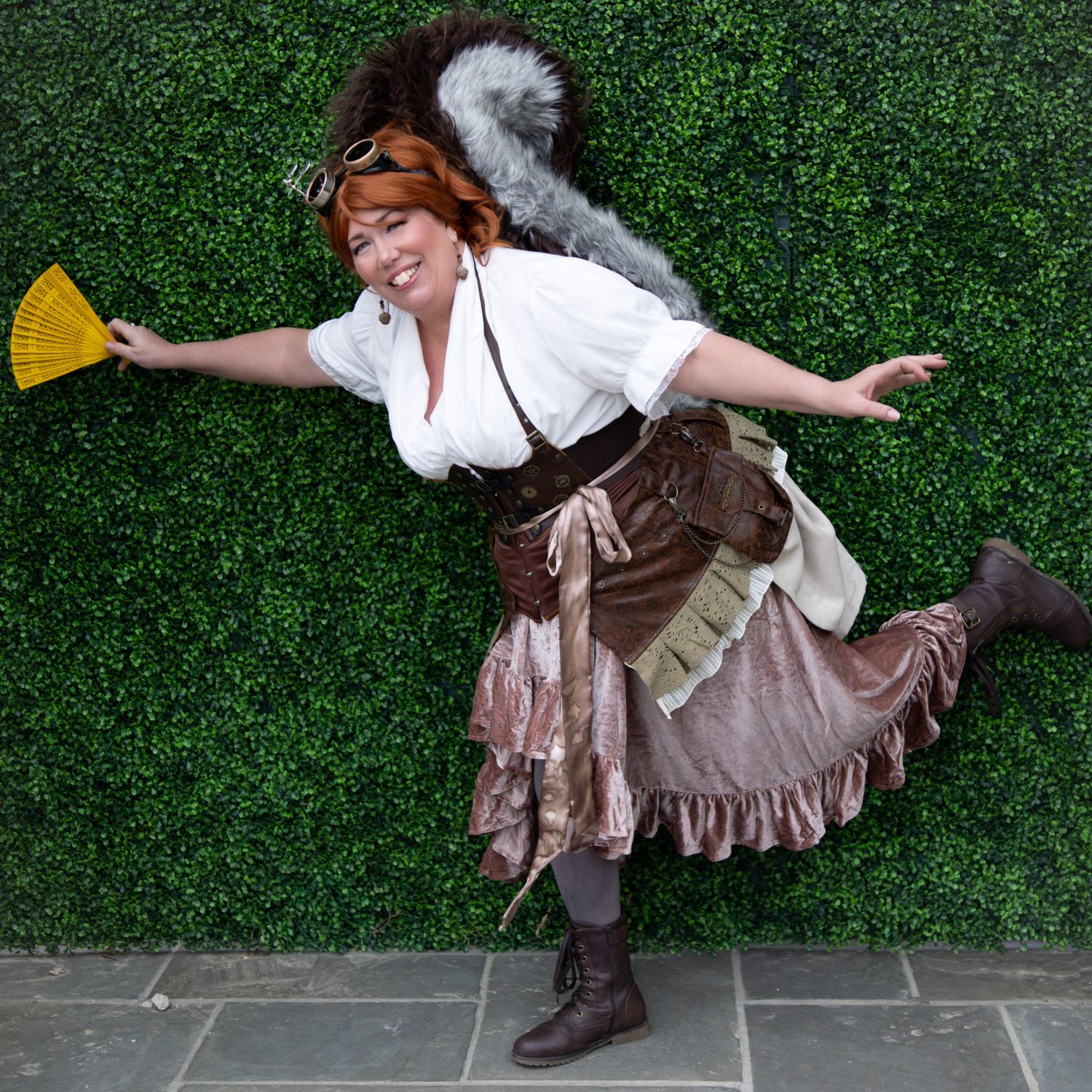 Woman in steampunk costume with squirrel tail, goggles, ruffled skirt, and boots poses playfully against green hedge wall.