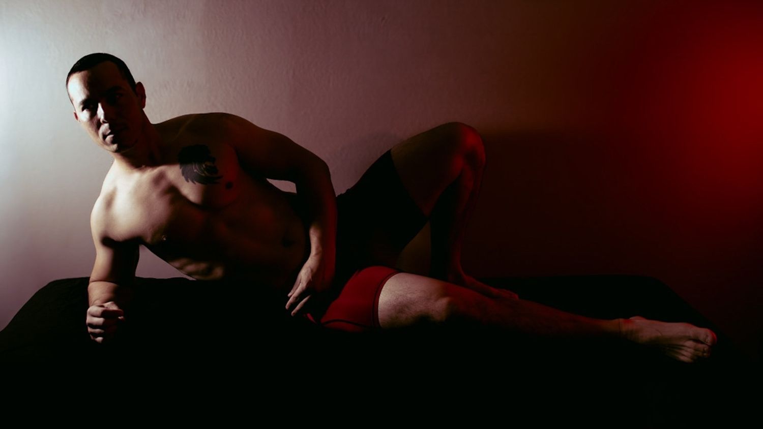 Shirtless athletic man in red shorts posing on a dark surface in dramatic red and white lighting.