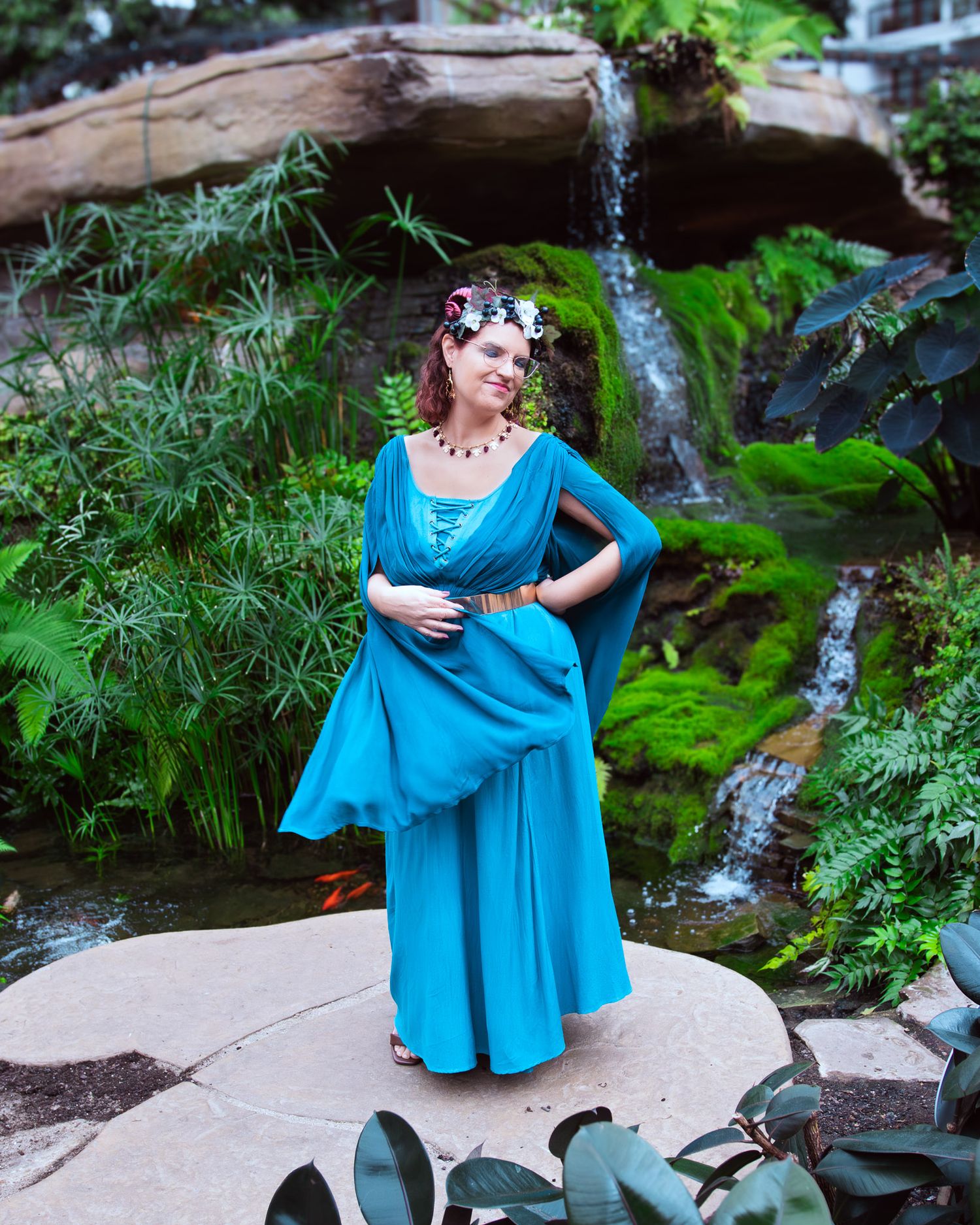 Woman in teal gown with floral crown poses near waterfall and lush greenery in enchanting garden setting.