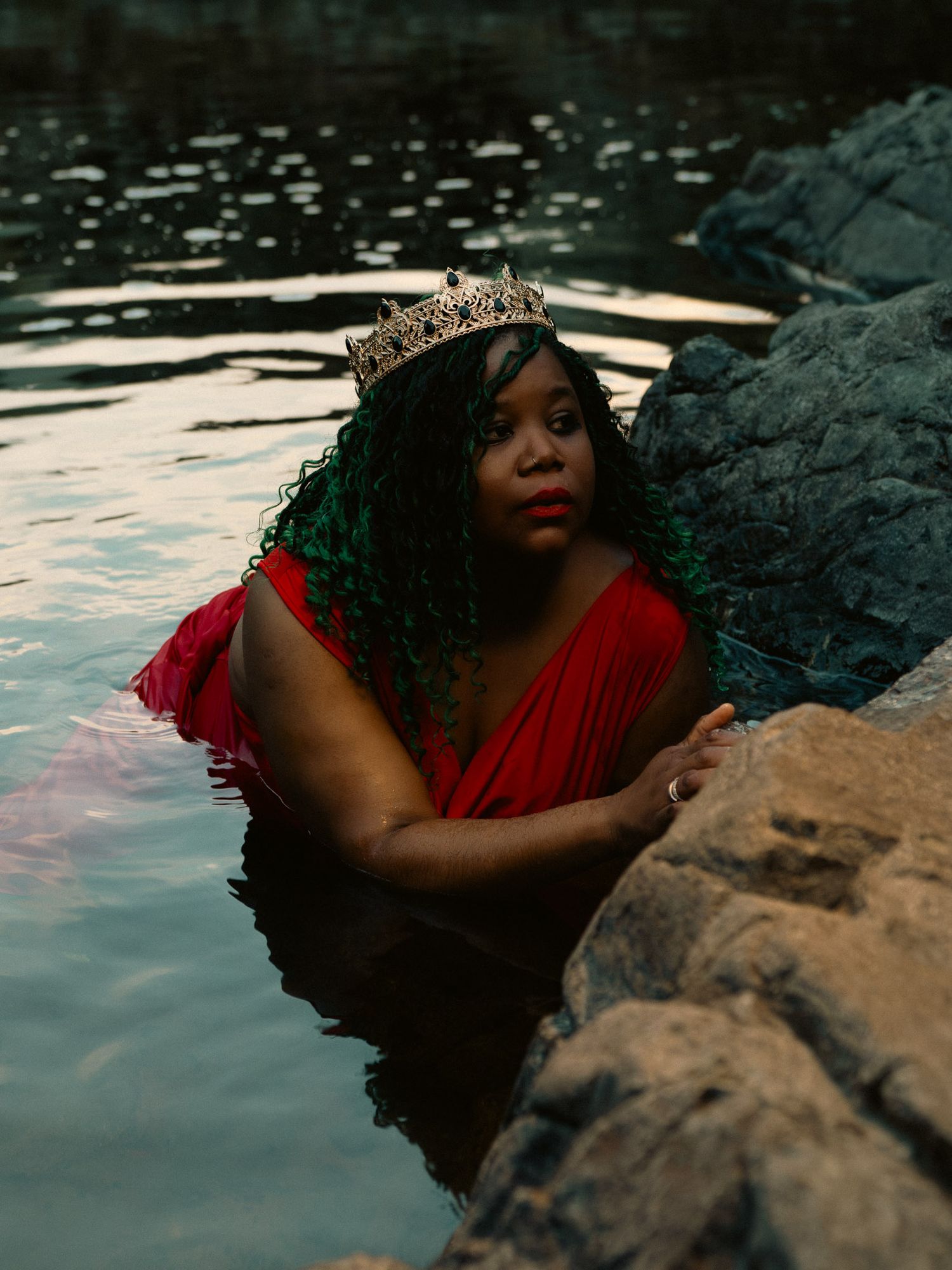 A Black woman wearing a red dress and jeweled crown poses regally in a rocky natural pool with green curly hair.
