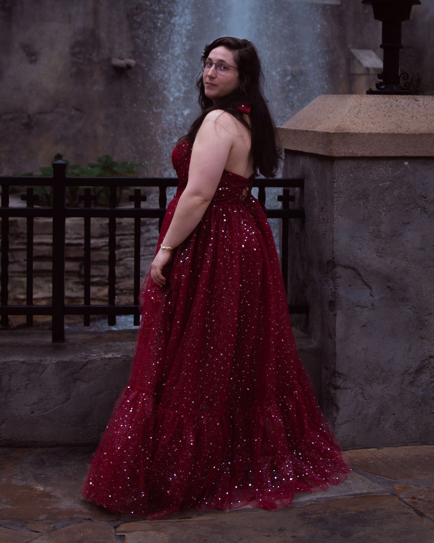 Woman in sparkling red sequin ball gown posing elegantly in front of a stone wall and fountain.