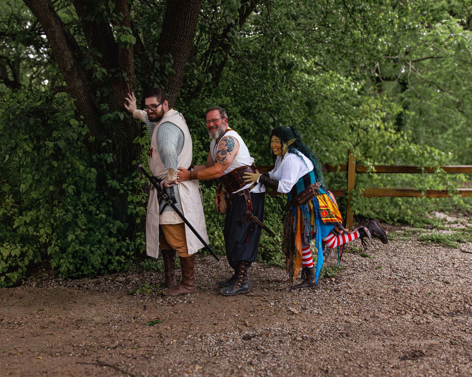 Three people in medieval Renaissance faire costumes pose outdoors near trees and a wooden fence on a dirt path.