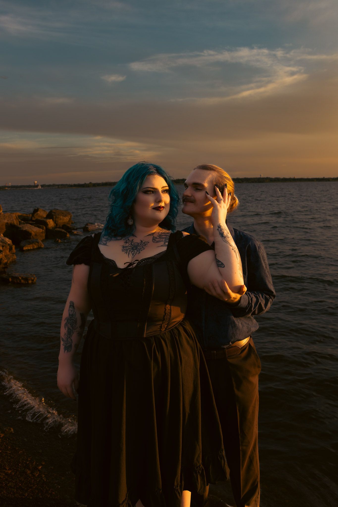 A tattooed couple with blue hair poses by rocky shoreline at golden sunset, wearing dark gothic-style outfits.