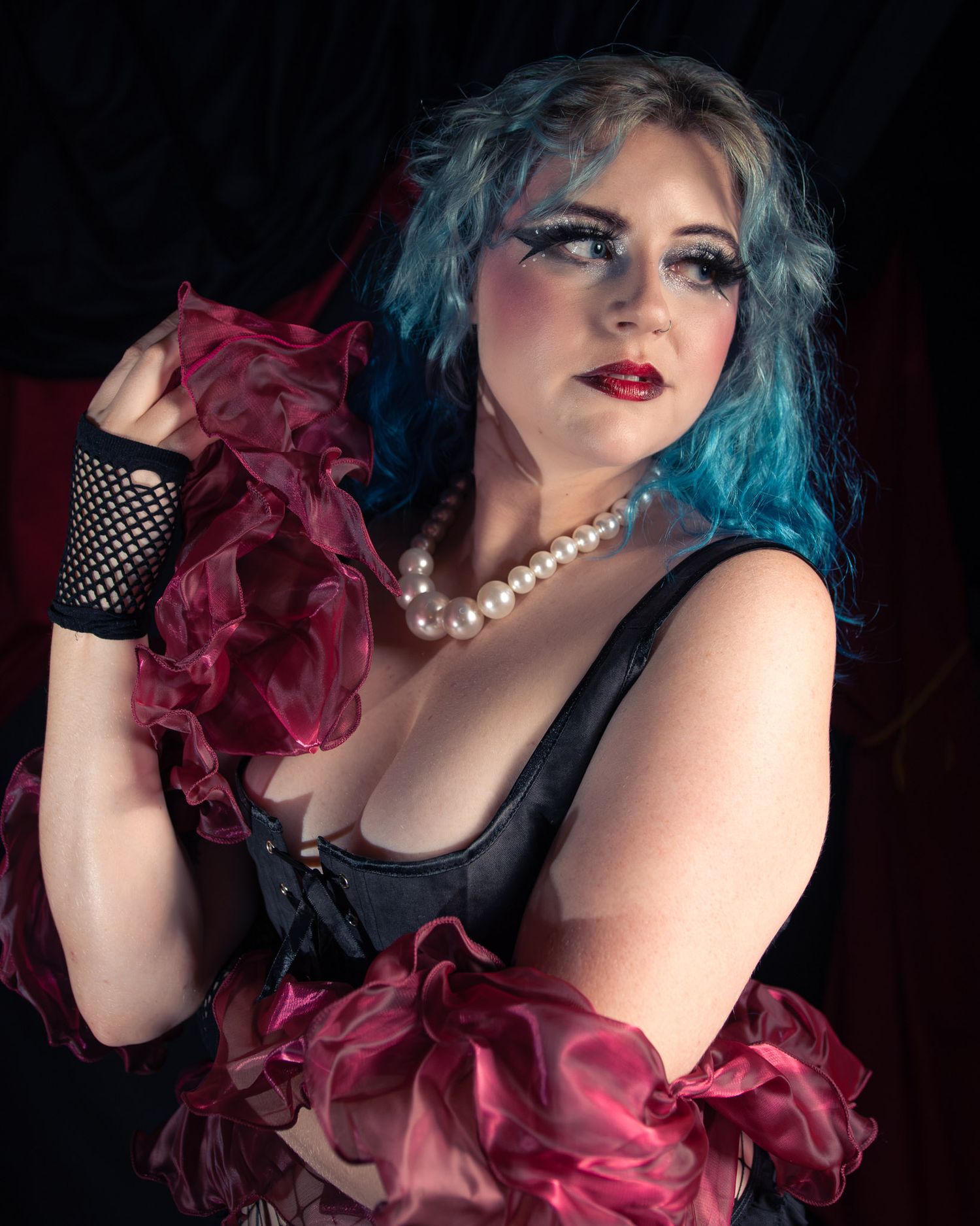Woman with blue hair in black corset and red ruffled boa, wearing pearl necklace and fishnet gloves.