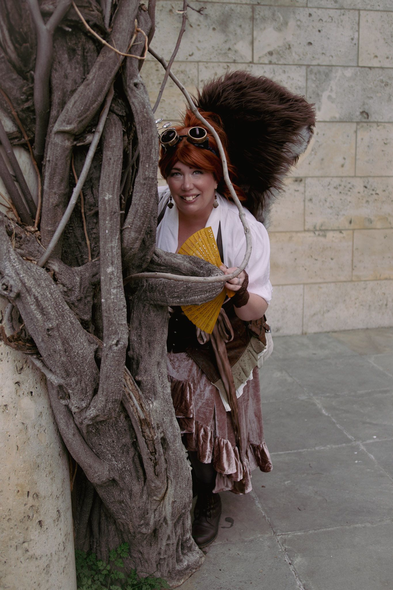 Woman in steampunk squirrel costume with fluffy tail and goggles poses beside a twisted tree trunk.