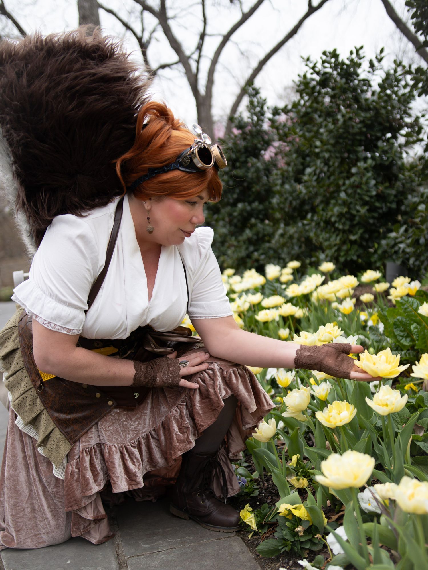 Woman in steampunk costume with goggles and squirrel tail crouches to touch yellow tulips in a garden.
