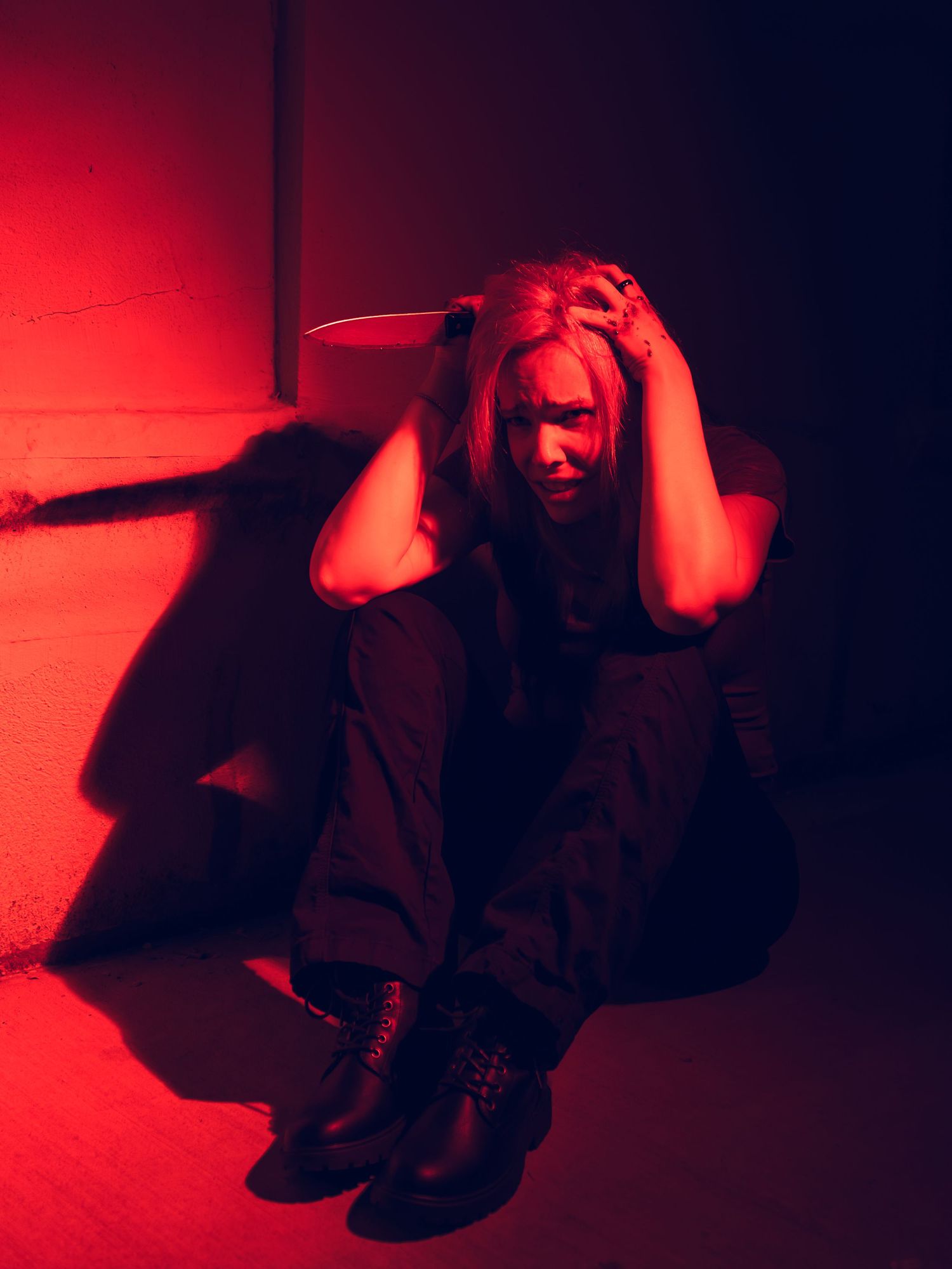 Woman sitting on the ground in red-lit alley, hands on head in distress and frustration, wearing dark clothing and boots, holding a knife.