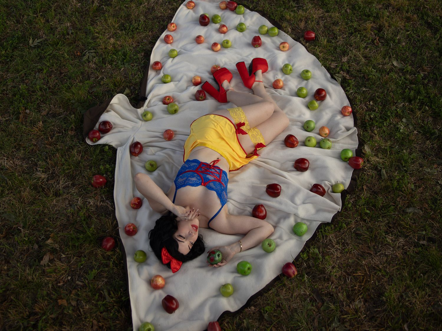 Woman in a Snow White-inspired costume lies on a white blanket on grass, surrounded by scattered red and green apples.