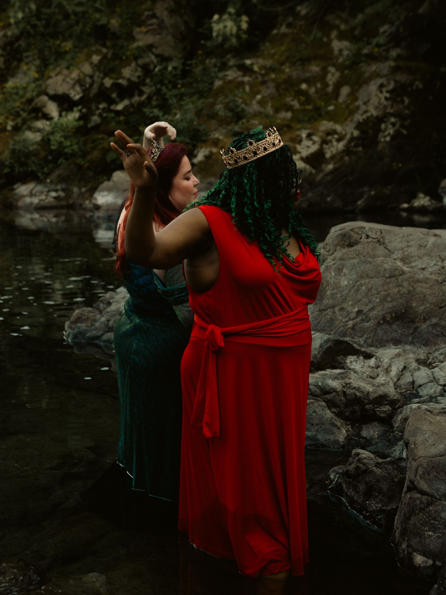 Two women in red and emerald green gowns with crowns stand by a rocky river in a fantasy-inspired outdoor photoshoot.
