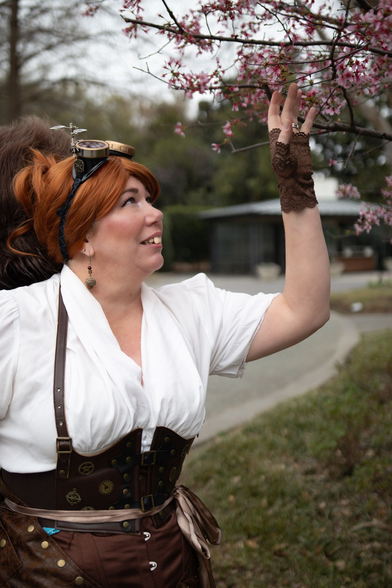A smiling redhead woman in steampunk costume with corset and goggles reaches up to touch pink cherry blossoms on a tree.