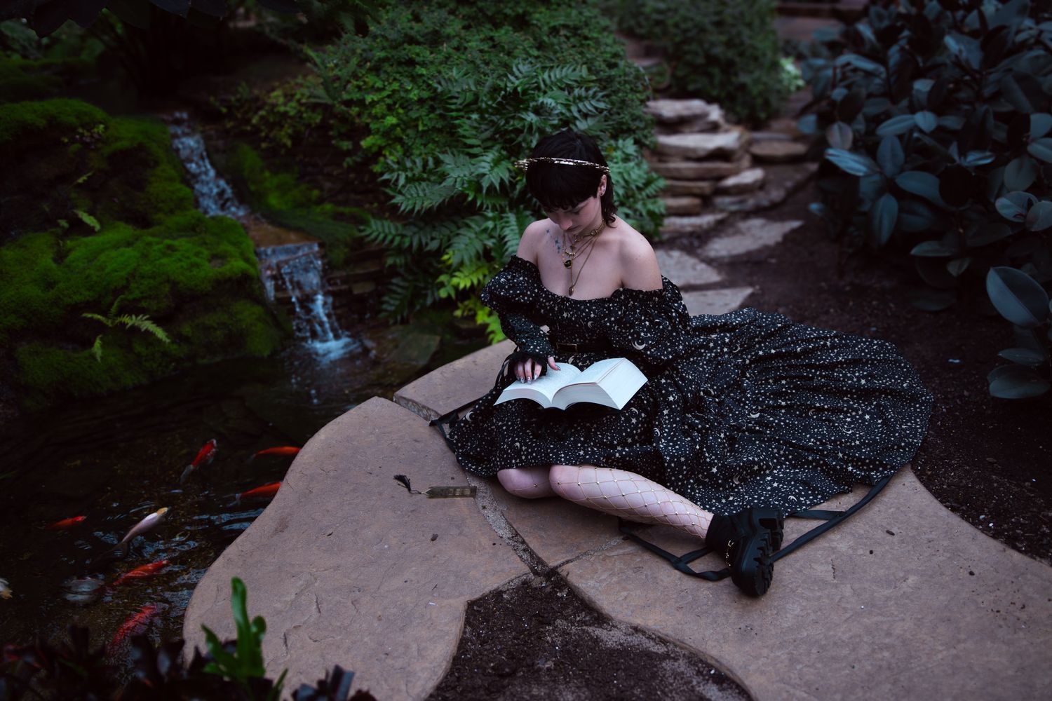 Woman in black starry gown reading a book beside a koi pond in a lush garden setting.