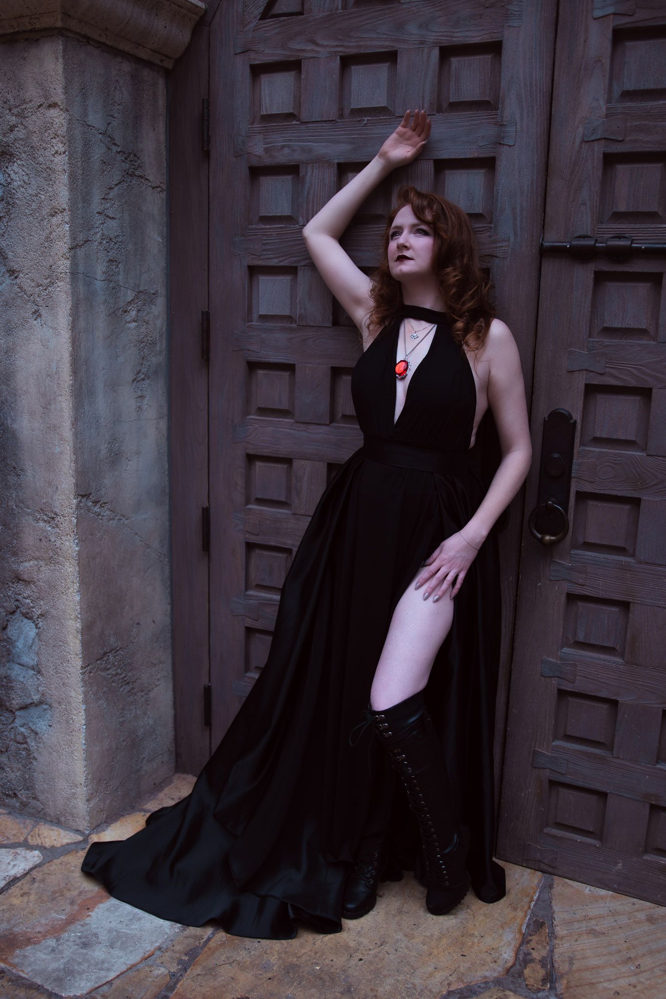 Woman in black halter gown with thigh slit and red necklace poses dramatically against ornate wooden doors.