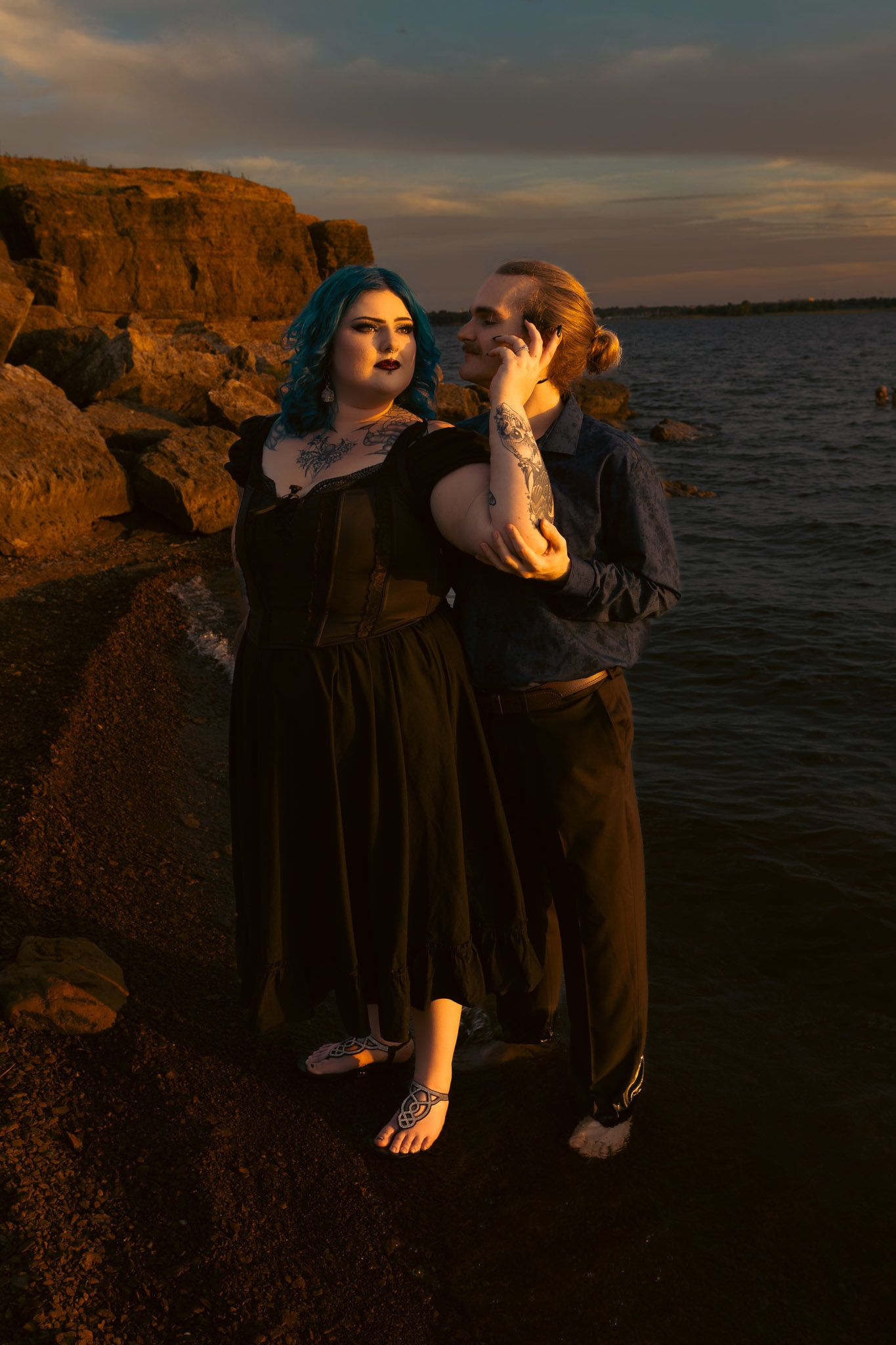 Couple with blue hair and tattoos posing at rocky shoreline during golden hour sunset.