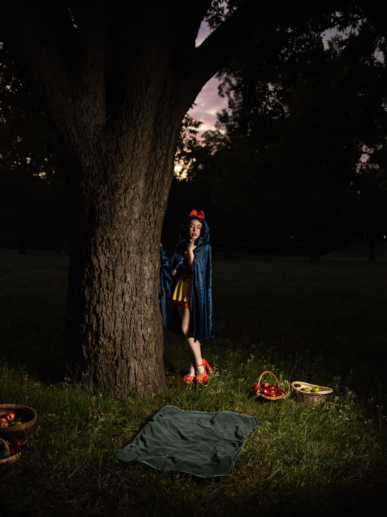 Person in a blue cape and red hair bow stands beside a large tree at dawn, with baskets of red apples and a green blanket on the grass.