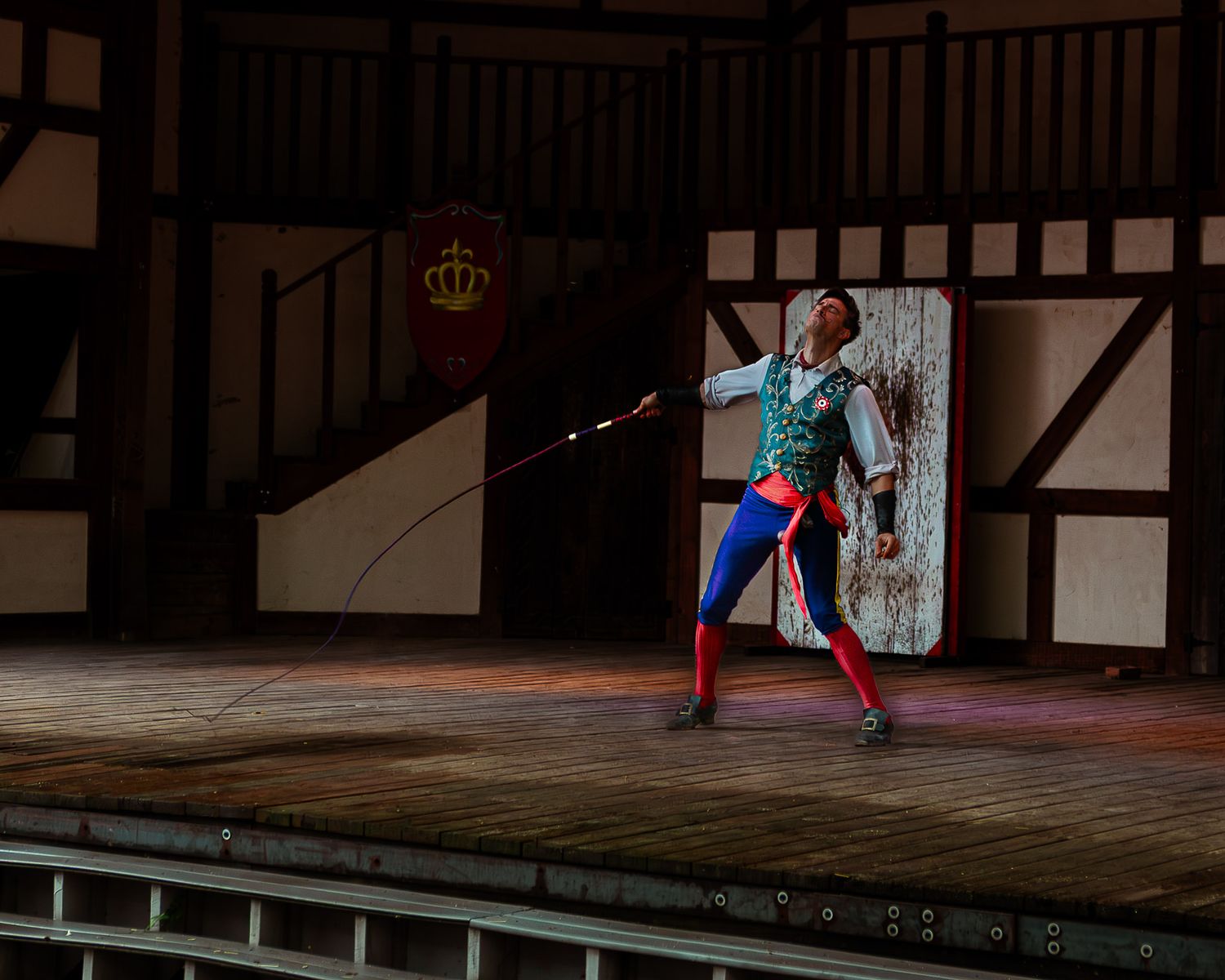 A performer in colorful medieval costume wielding a whip on a wooden stage during a theatrical Renaissance faire performance.