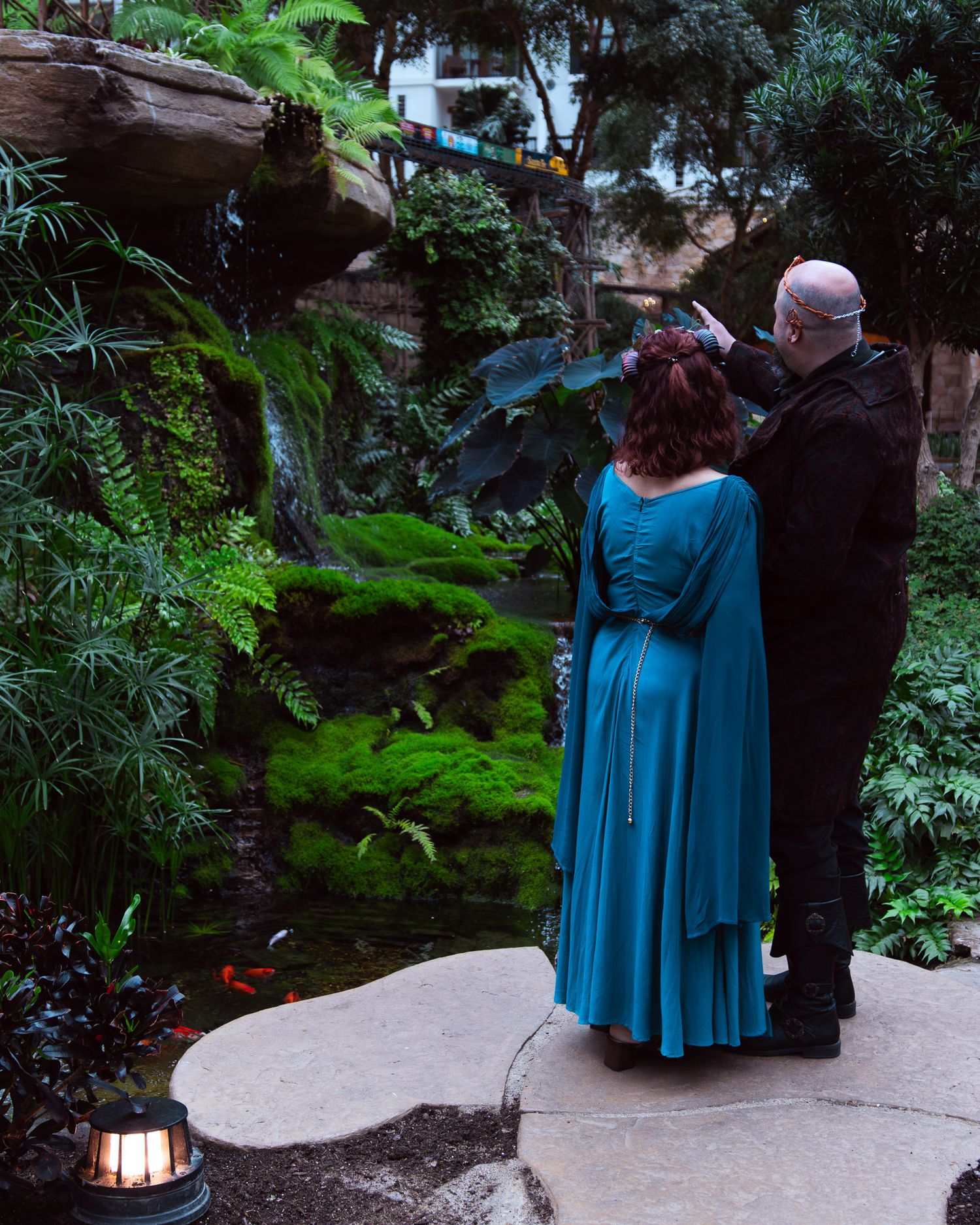 Couple in teal dress and black outfit stand in lush garden with ferns, moss, and koi pond.