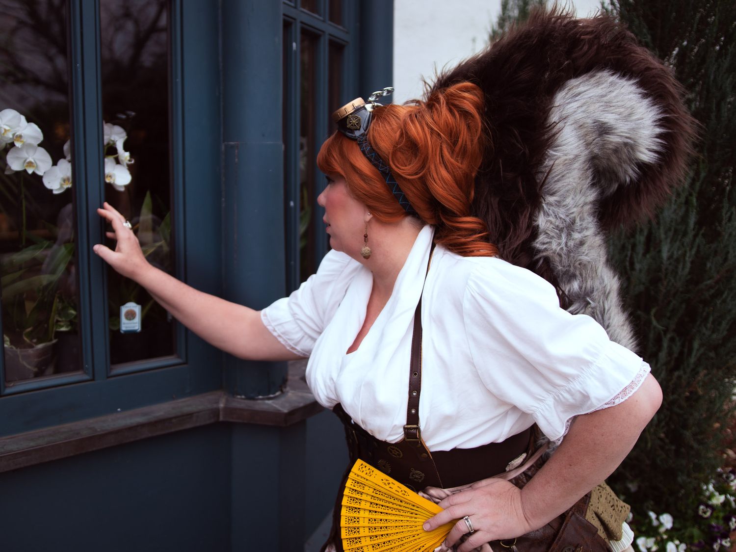 Woman in steampunk costume with red hair, white blouse, brown corset, yellow fan, and squirrel tail, pointing at window.