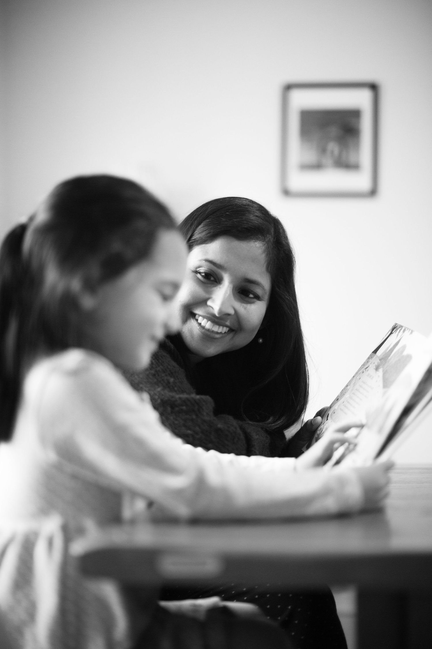 A mother is smiling as her daughter is reading a book at a table. Motherhood and childhood photo session by The Mother's Eye in Greenwich.
