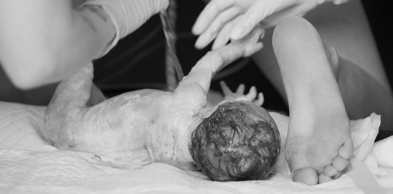 A newborn seconds after birth laying next to the foot of his mum on all fours. Home birth photography East Dulwich, South East London.