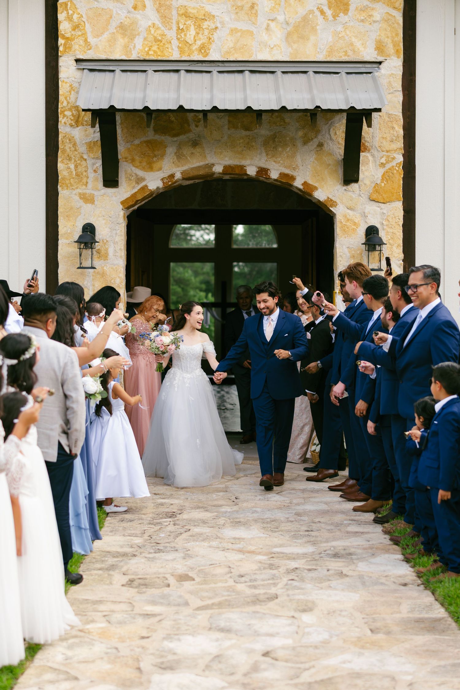Newlyweds exit stone chapel through cheering guests in grand wedding recessional celebration.