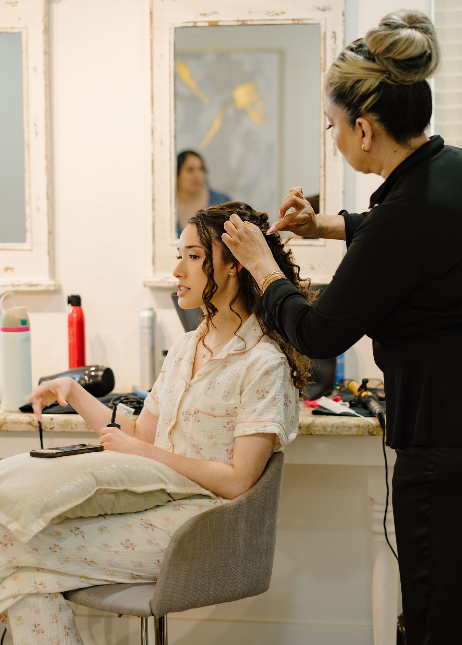 A hairstylist curling a young woman's hair in a bright salon while she sits in a styling chair.