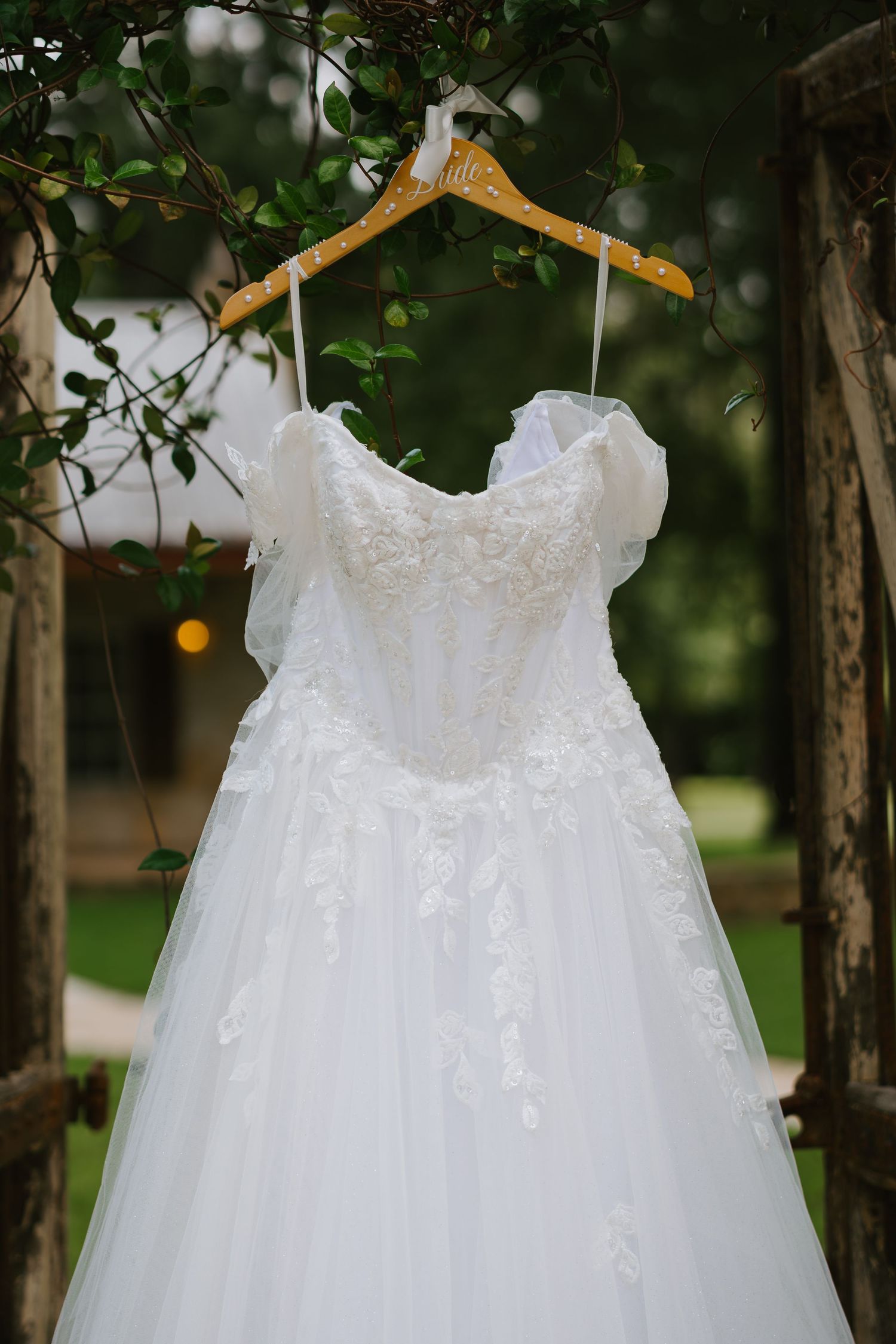 Elegant white lace ball gown wedding dress hanging on a wooden hanger outdoors among green foliage.