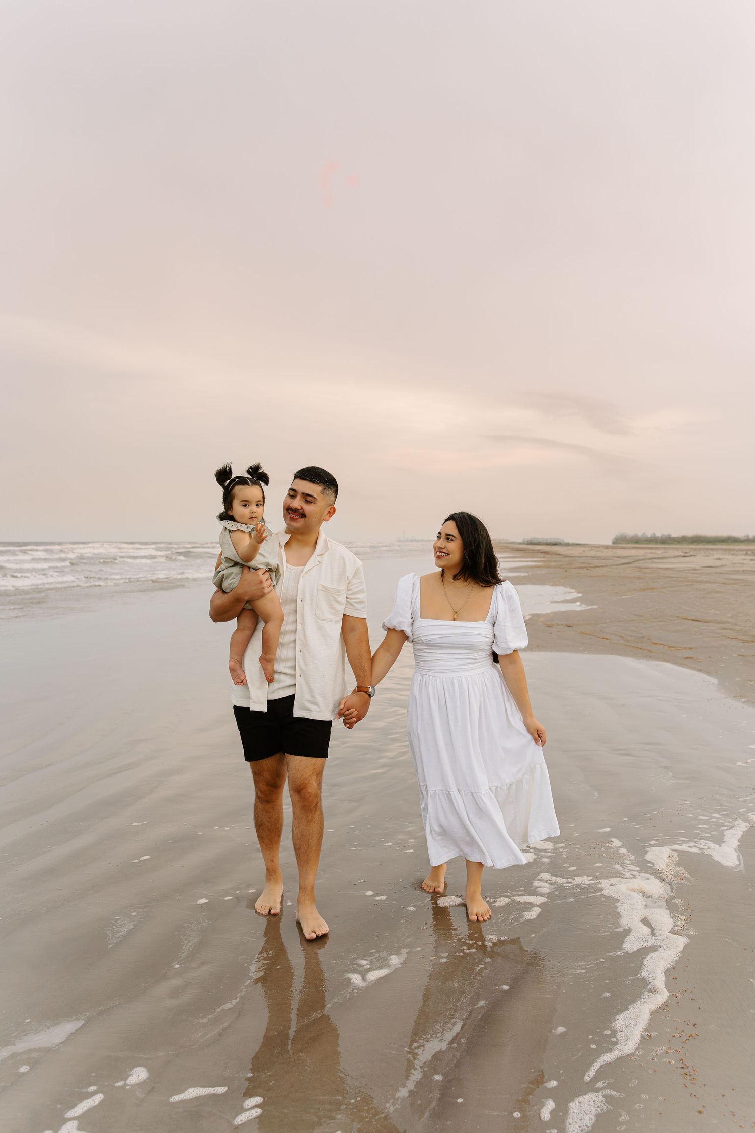 Family of three walks on beach at sunset, parents holding hands, father carrying toddler, wearing white outfits.