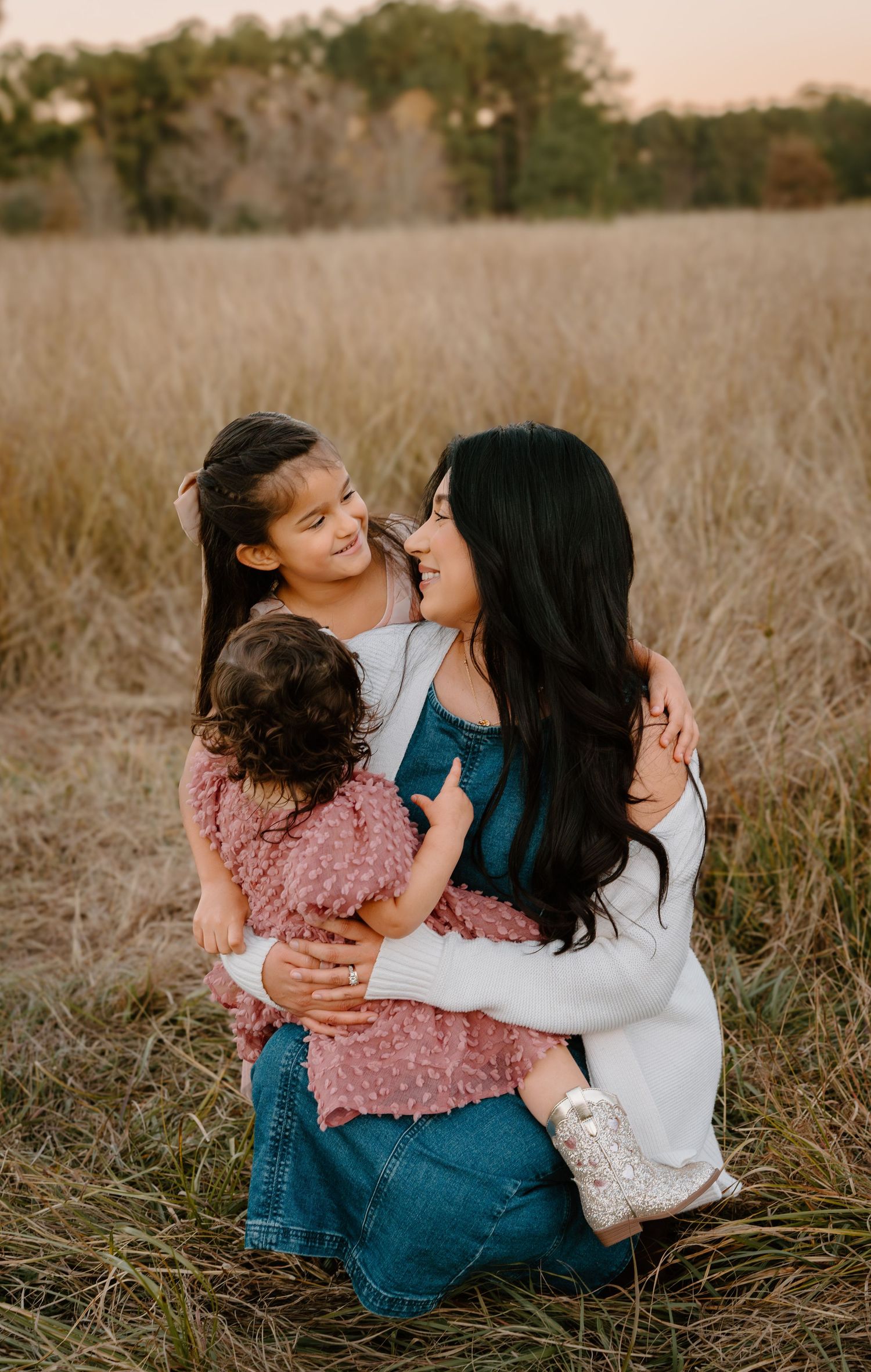 A mother sits in a golden field holding two young daughters, smiling warmly in a tender outdoor family portrait.