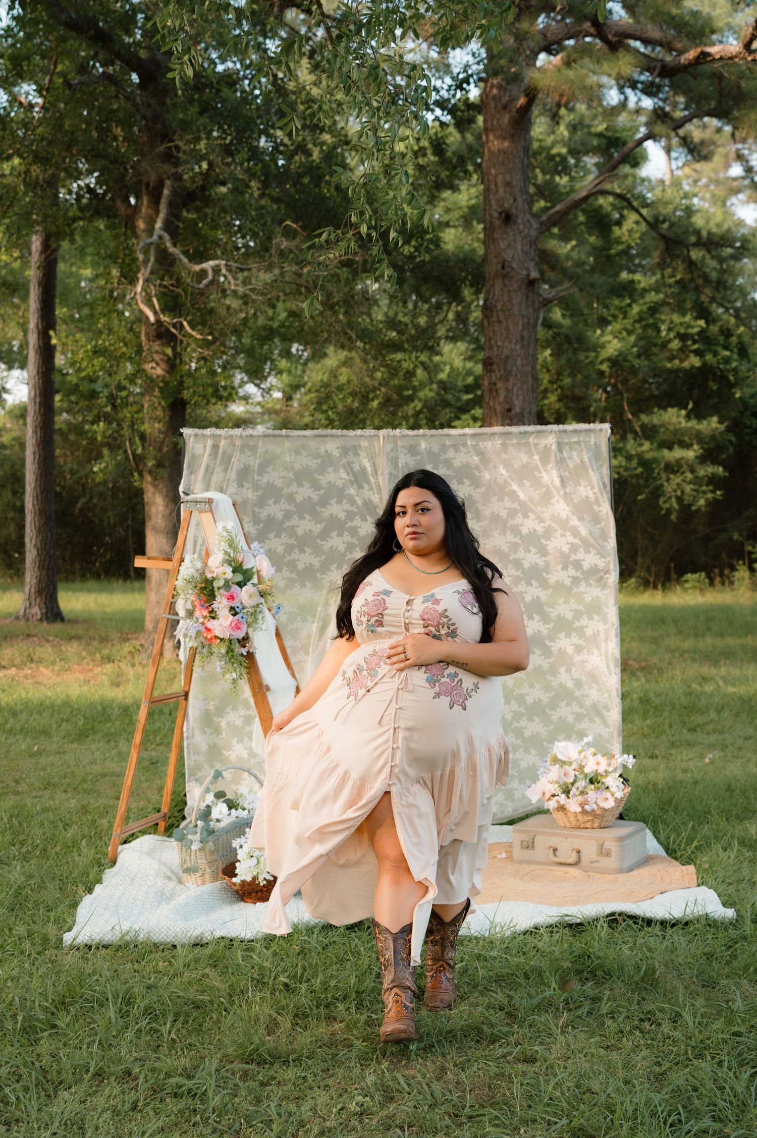 Woman in floral dress and boots poses outdoors at boho-styled photo shoot with lace backdrop and floral arrangements.