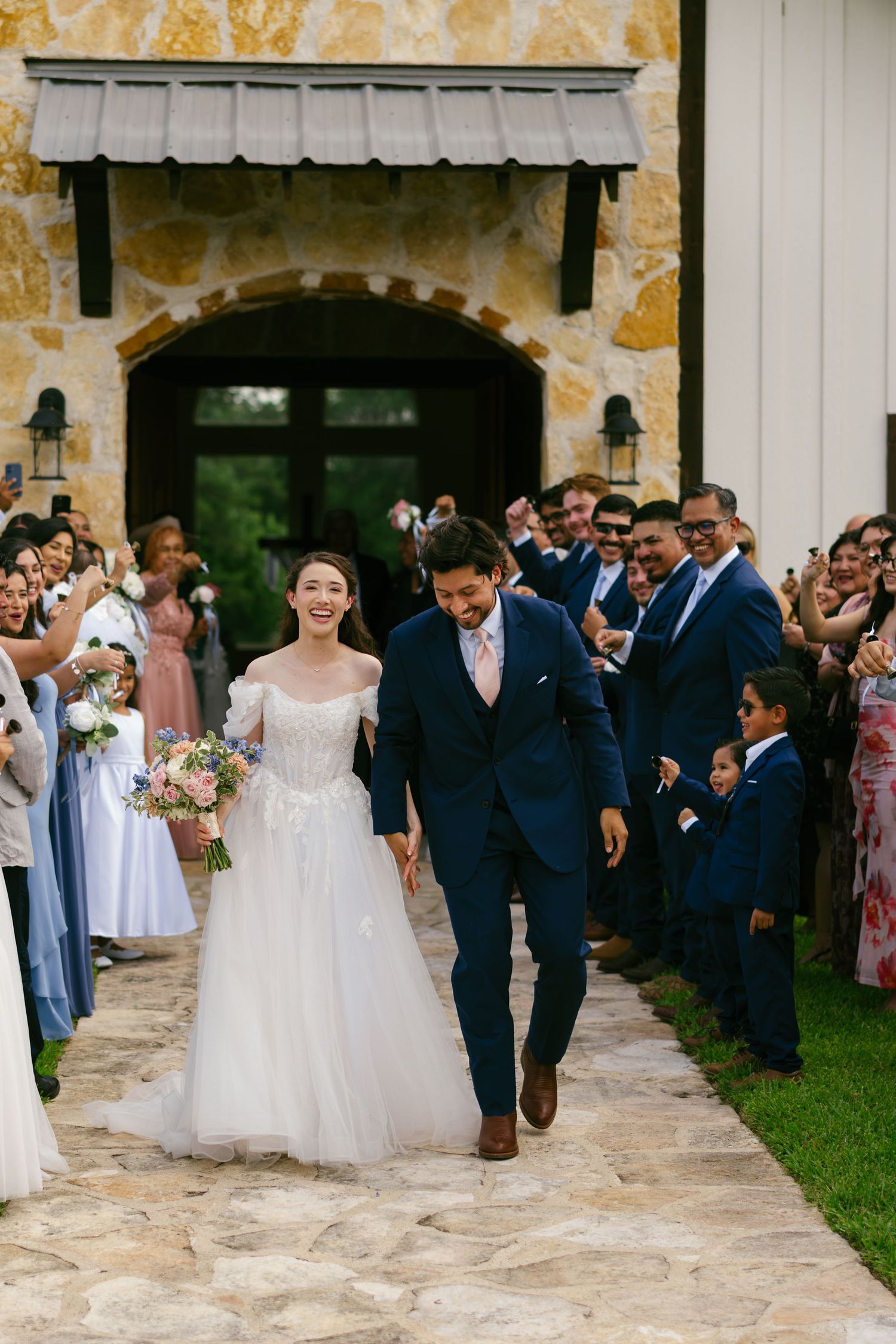 Joyful newlyweds walk down stone path as guests celebrate their wedding ceremony exit outside a rustic chapel.
