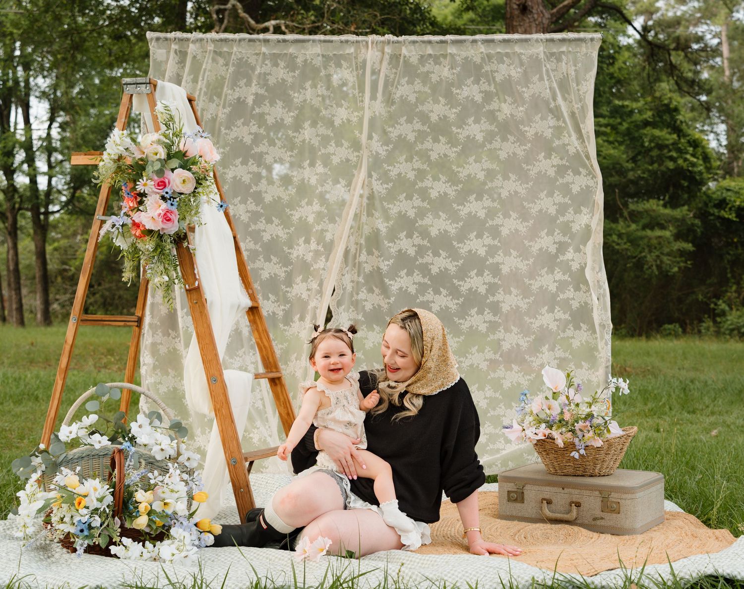 Woman and baby girl pose outdoors by lace backdrop with floral arrangements and vintage suitcase on blanket.