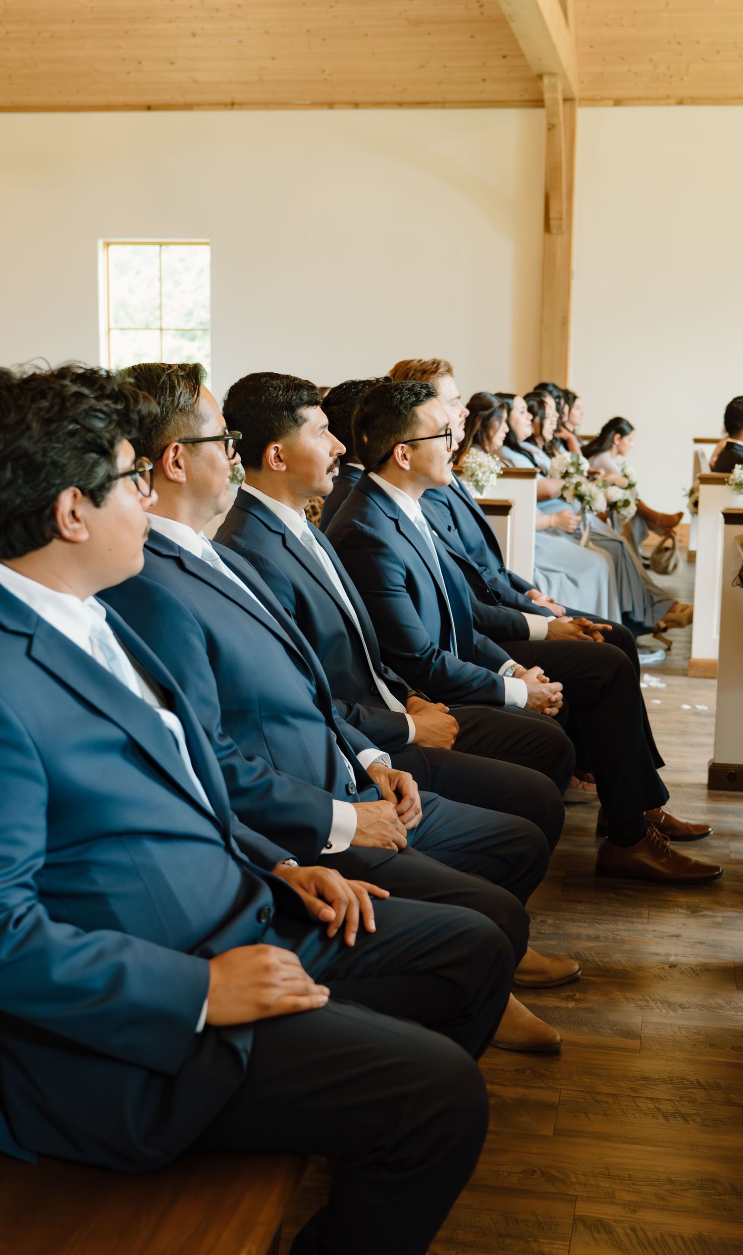 Groomsmen in matching navy suits seated in wooden chapel pews during wedding ceremony.