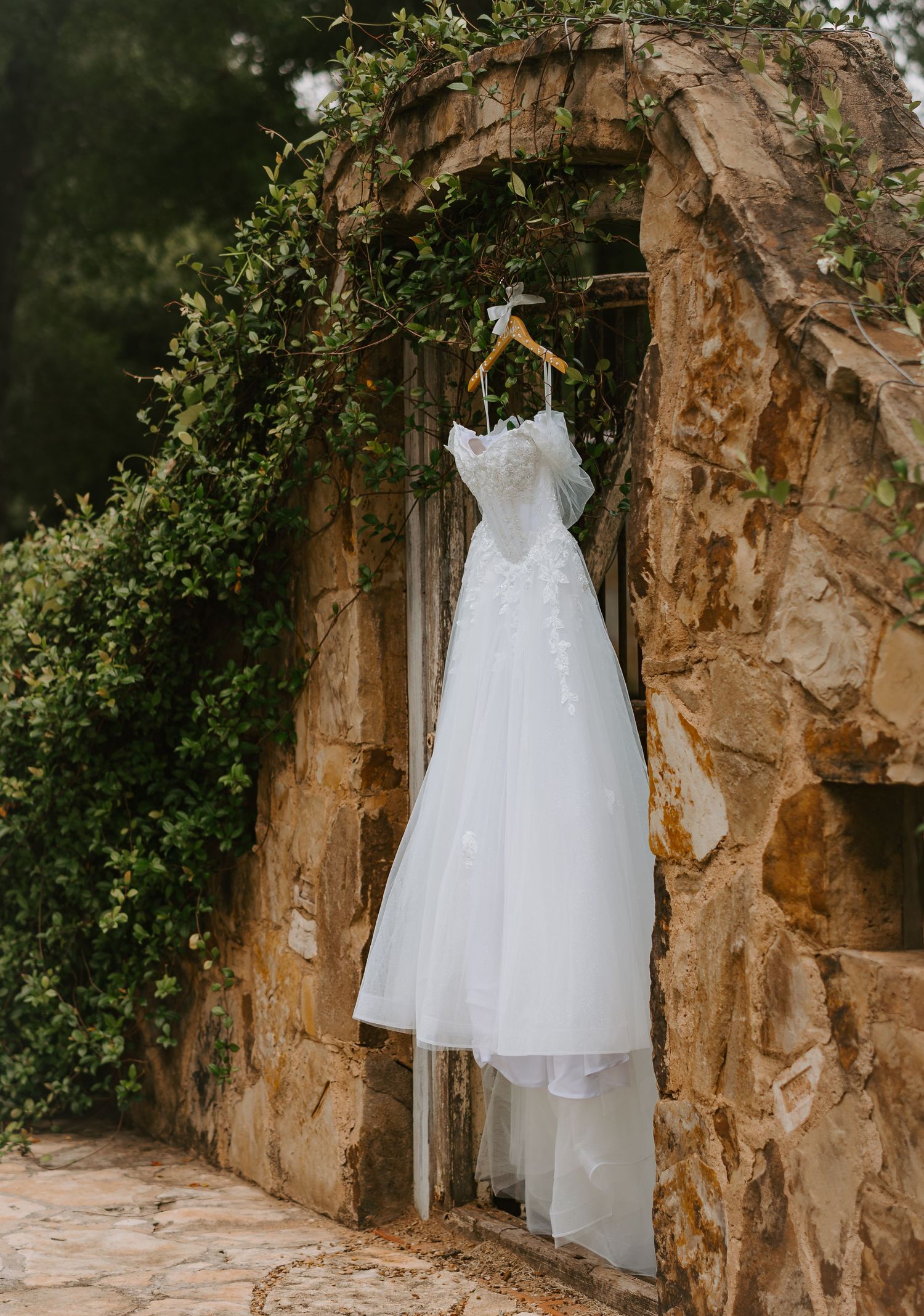 A white lace A-line wedding dress hanging on a wooden hanger in a rustic stone archway with greenery.