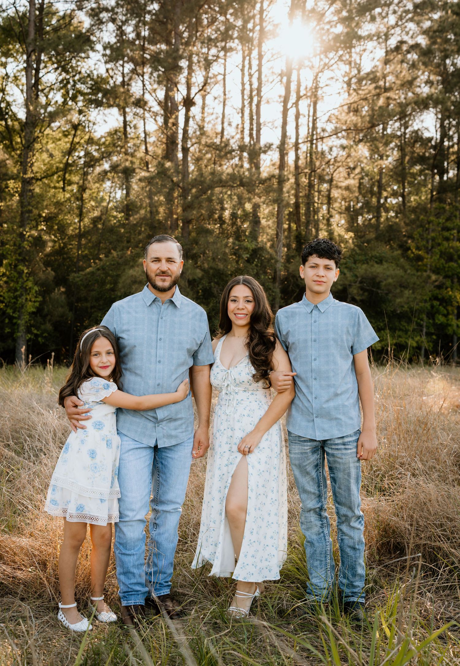 Family of four posing in a sunlit meadow with tall pine trees, wearing coordinated blue and white outfits.