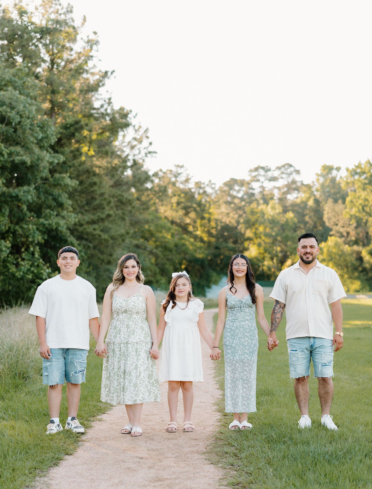 Family of five walking hand-in-hand on a sunlit path surrounded by lush green trees, dressed in coordinated outfits.