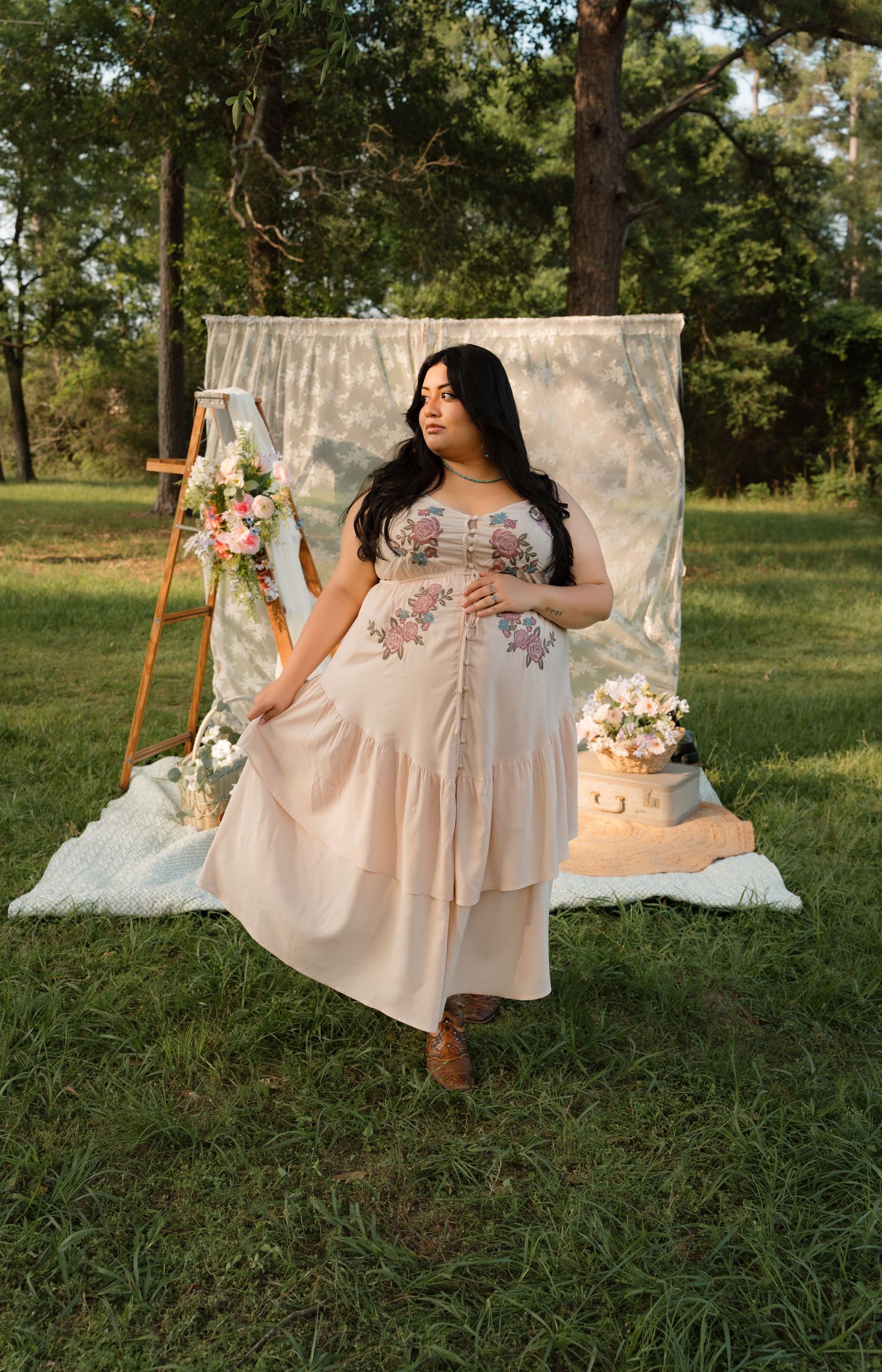 Woman in floral embroidered blush maxi dress posing outdoors with boho backdrop, easel, and pink flowers on grass.