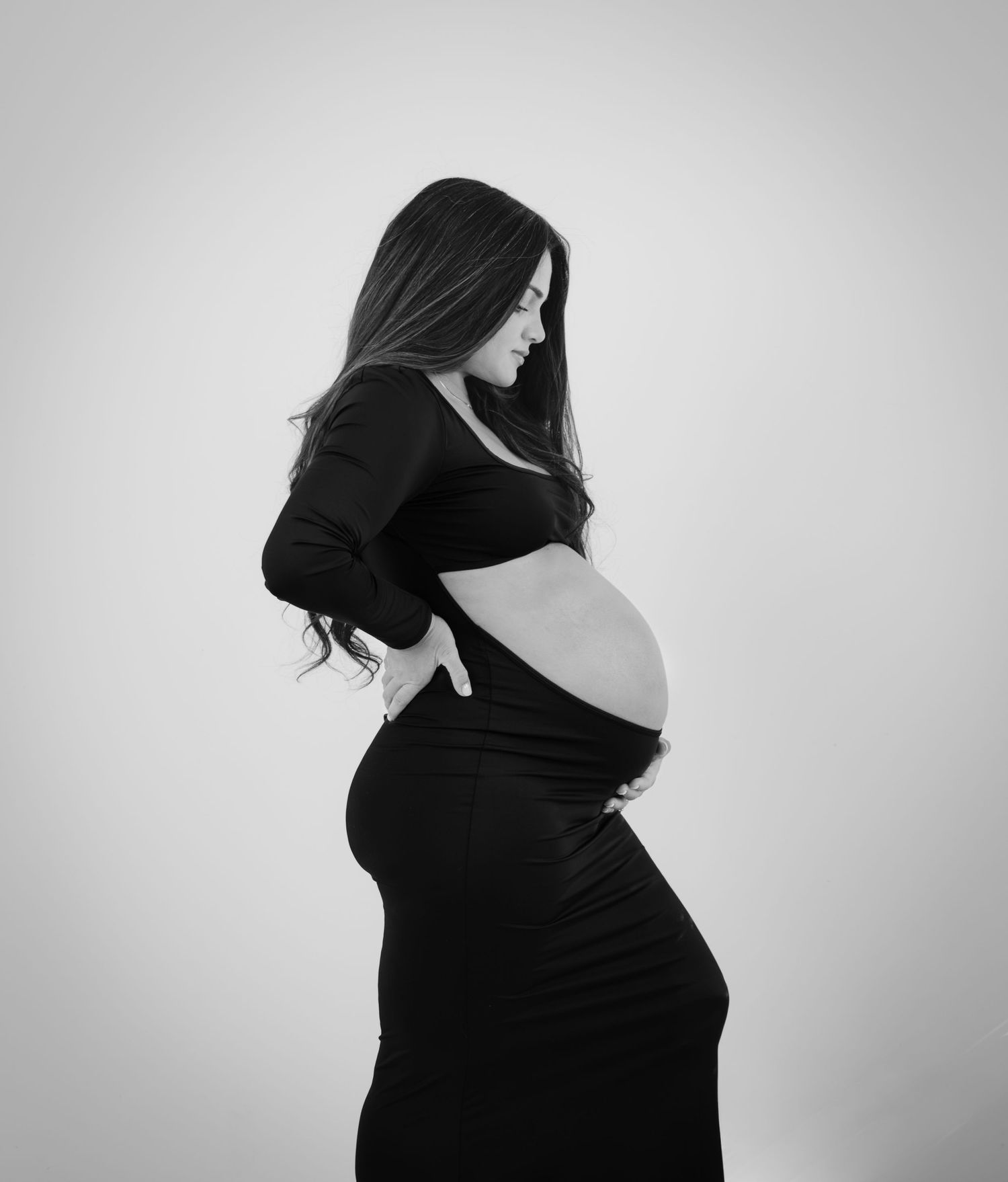 Black and white maternity photo of a pregnant woman in a black dress, elegantly posing sideways to showcase her baby bump.