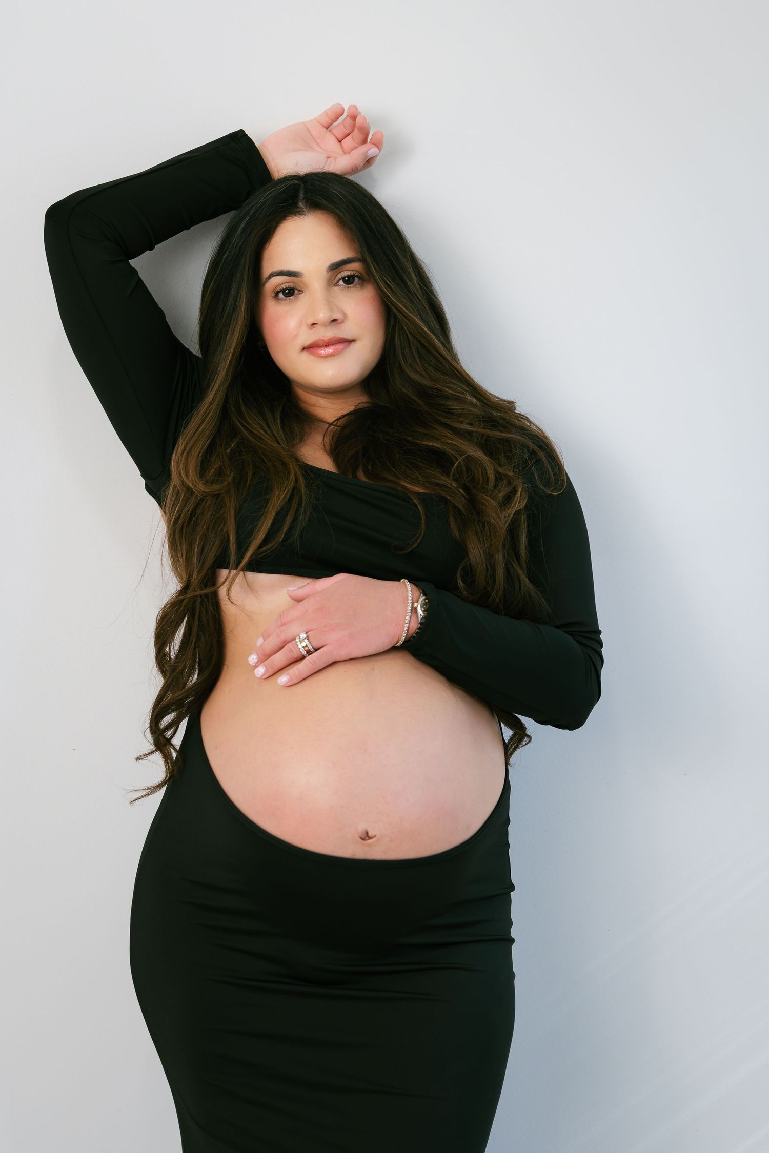 Pregnant woman in black outfit cradling baby bump, long dark hair, posing against white background.