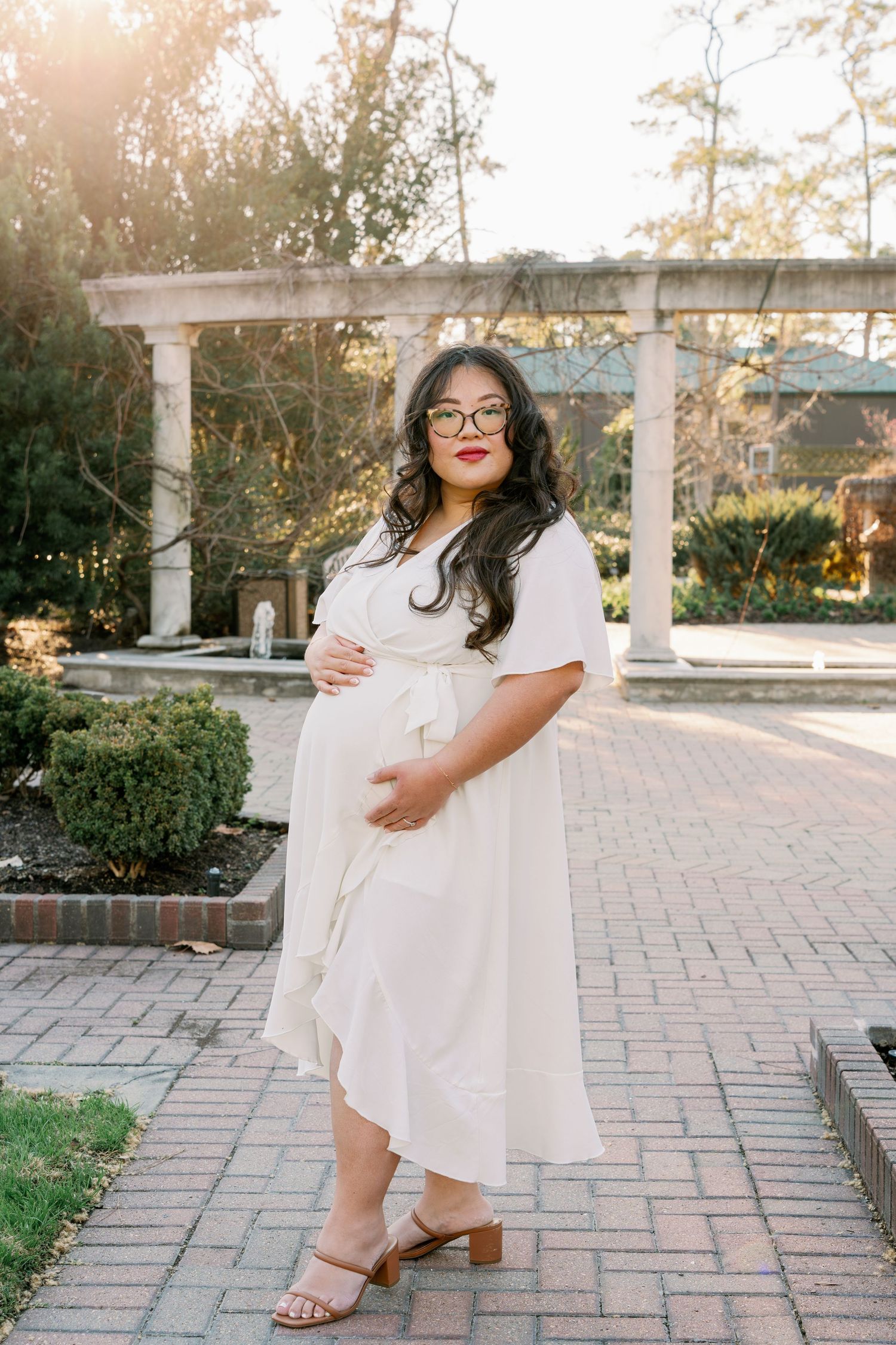 Pregnant woman in white wrap dress and glasses poses in a sunlit garden with brick pathway and stone columns.