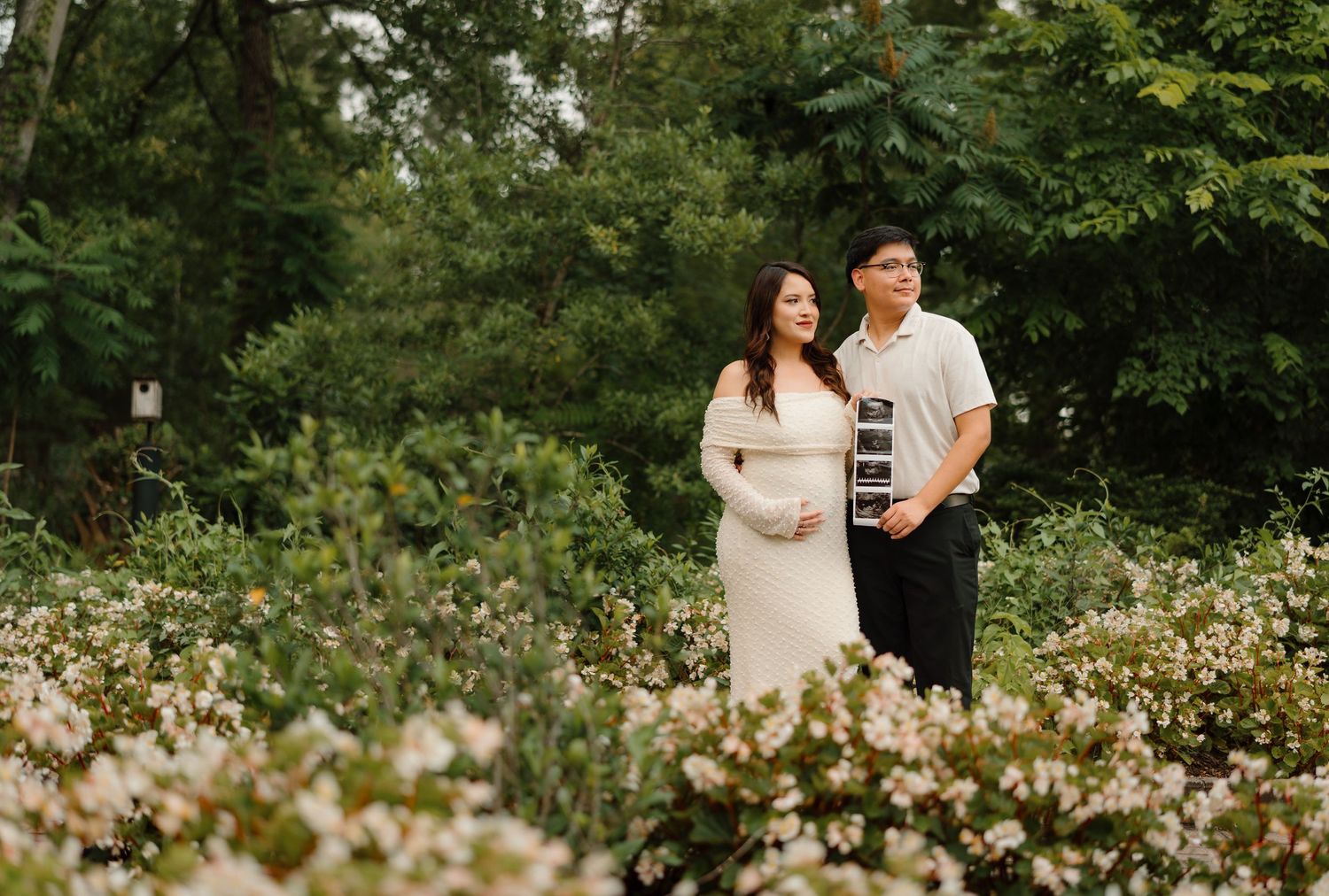 Couple standing in a lush garden with white flowers, woman in white dress holding ultrasound photos during maternity shoot.