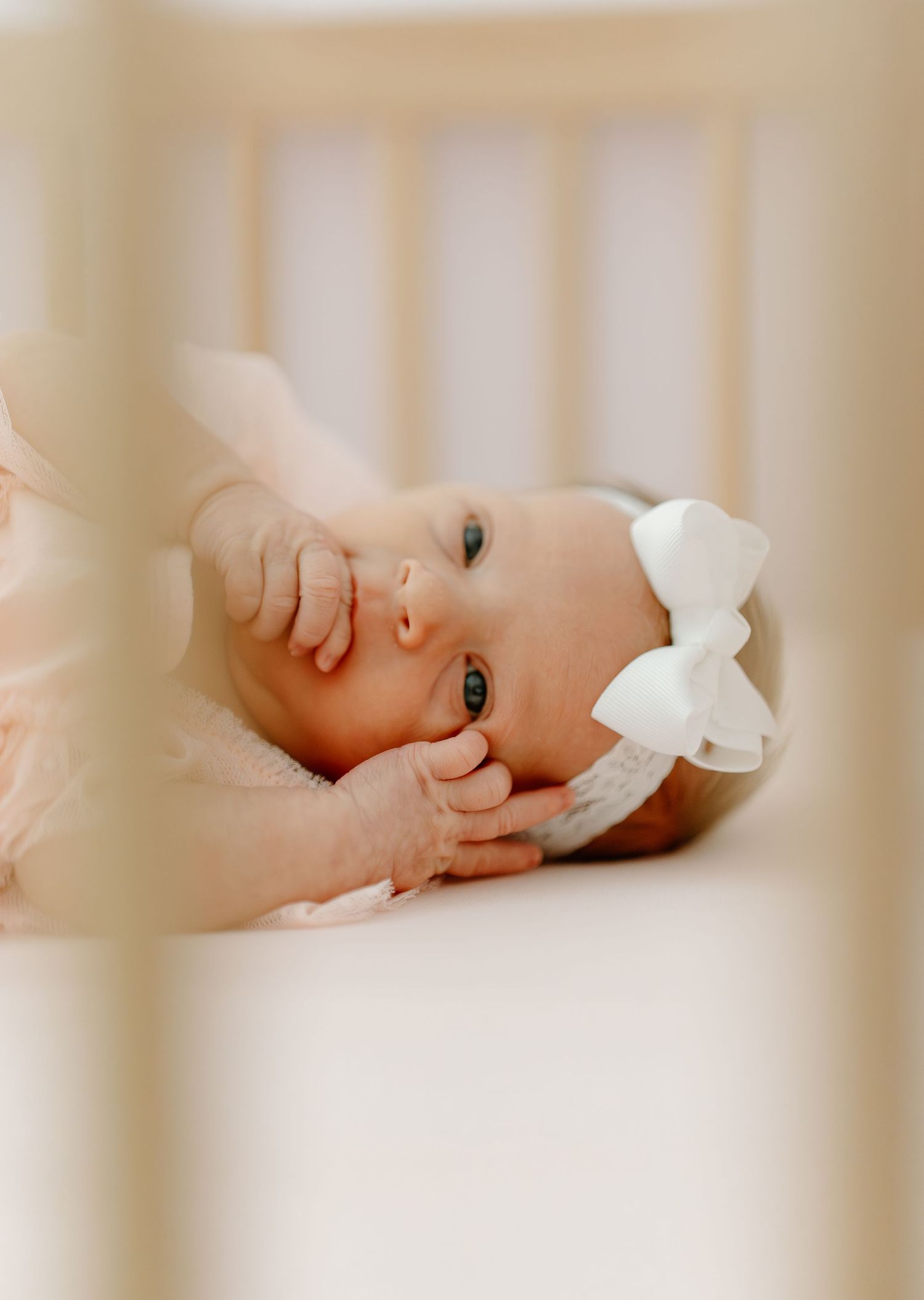 A baby girl with blue eyes wearing a white bow headband lies peacefully in a white crib.