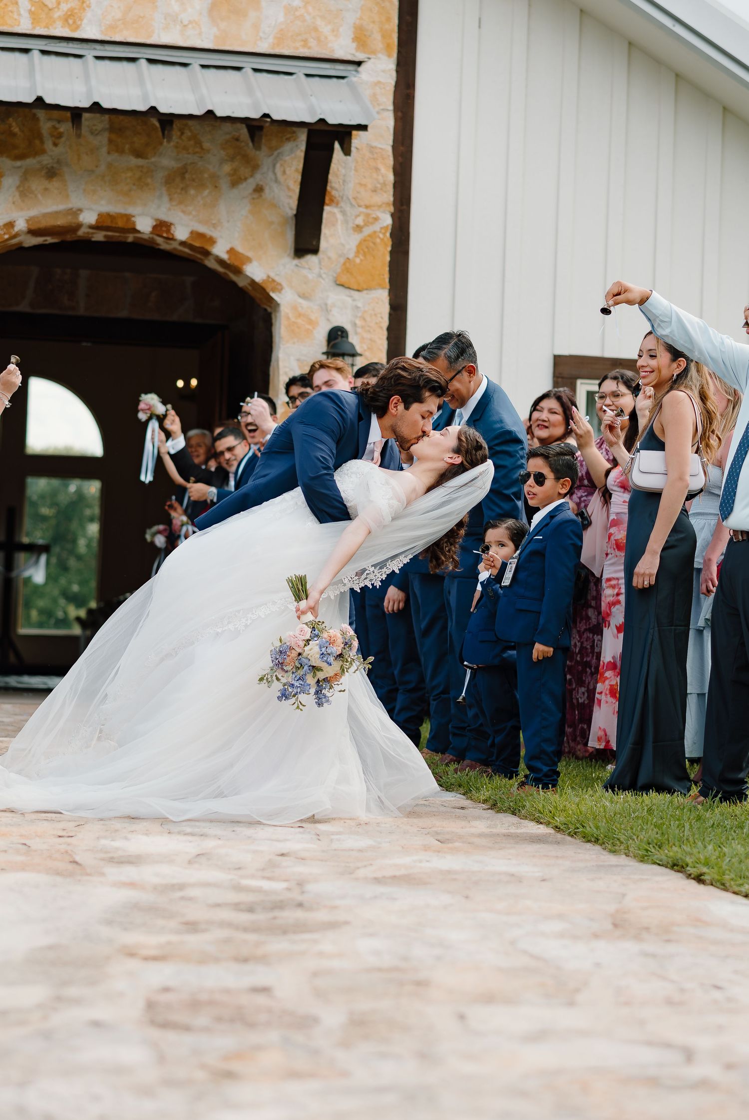 Groom dips bride for a kiss outside rustic stone wedding venue as bridal party cheers in navy blue attire.