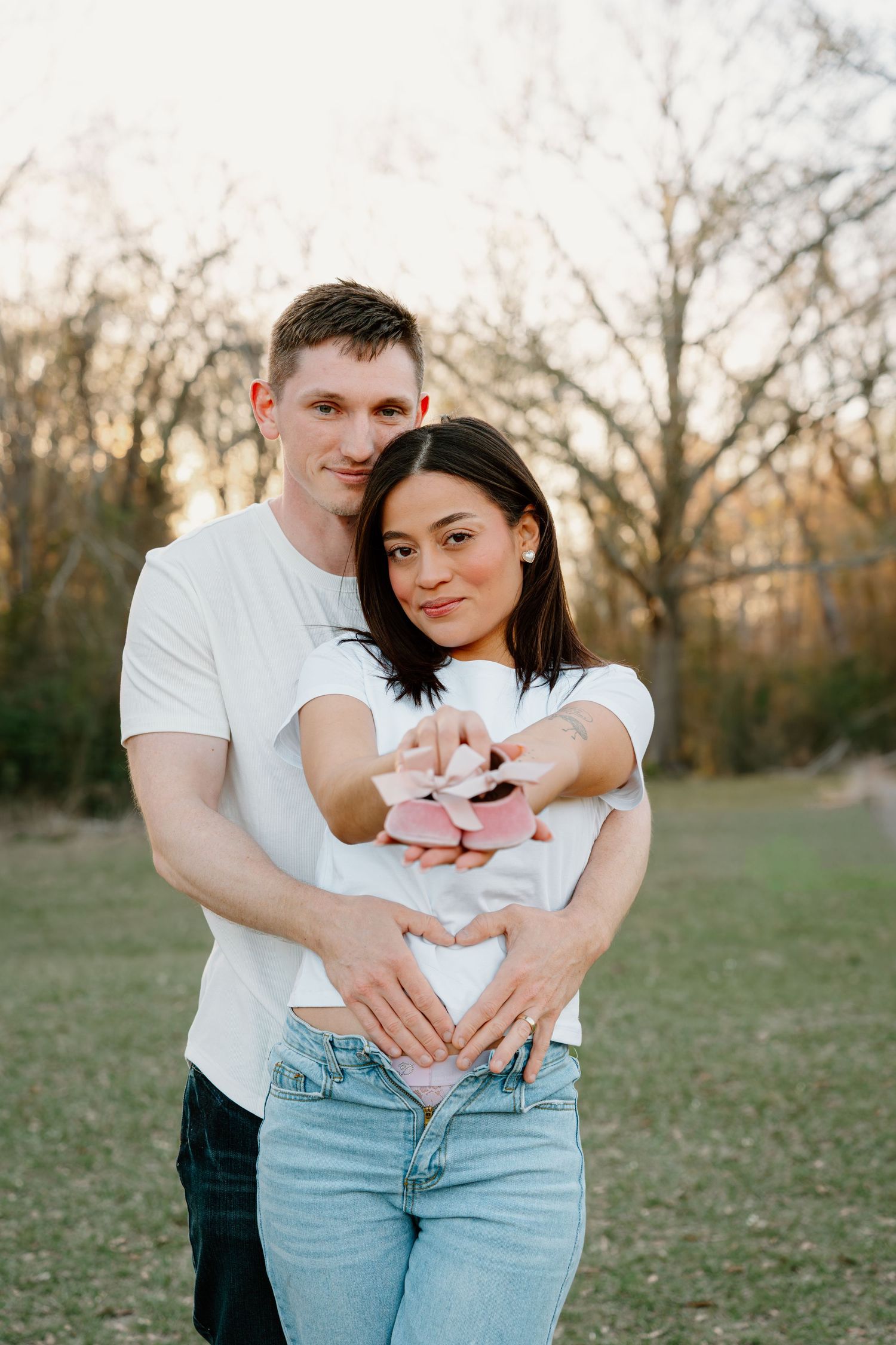 Expecting couple holding pink baby shoes forming a heart shape over baby bump outdoors.