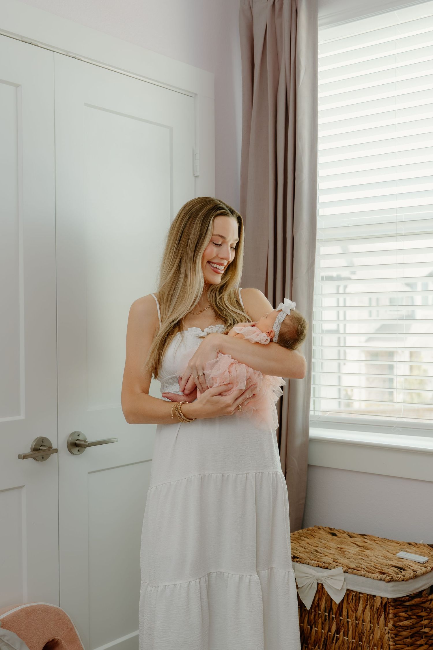 A smiling blonde woman in a white dress holds a newborn baby in a pink outfit near a bright window.