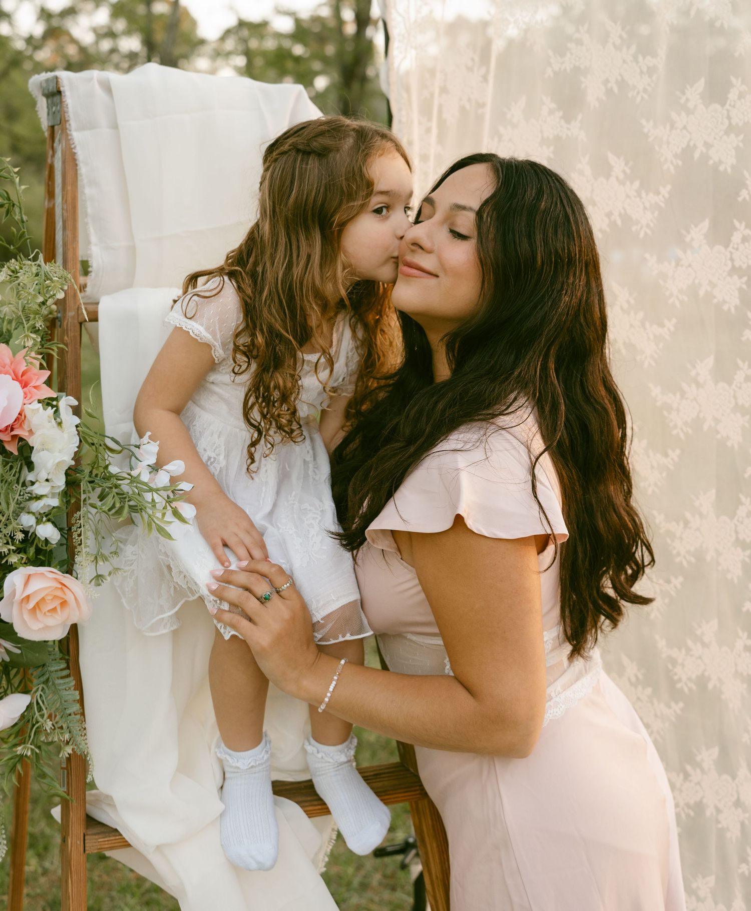 Young girl kissing mother on cheek outdoors near floral arrangement.