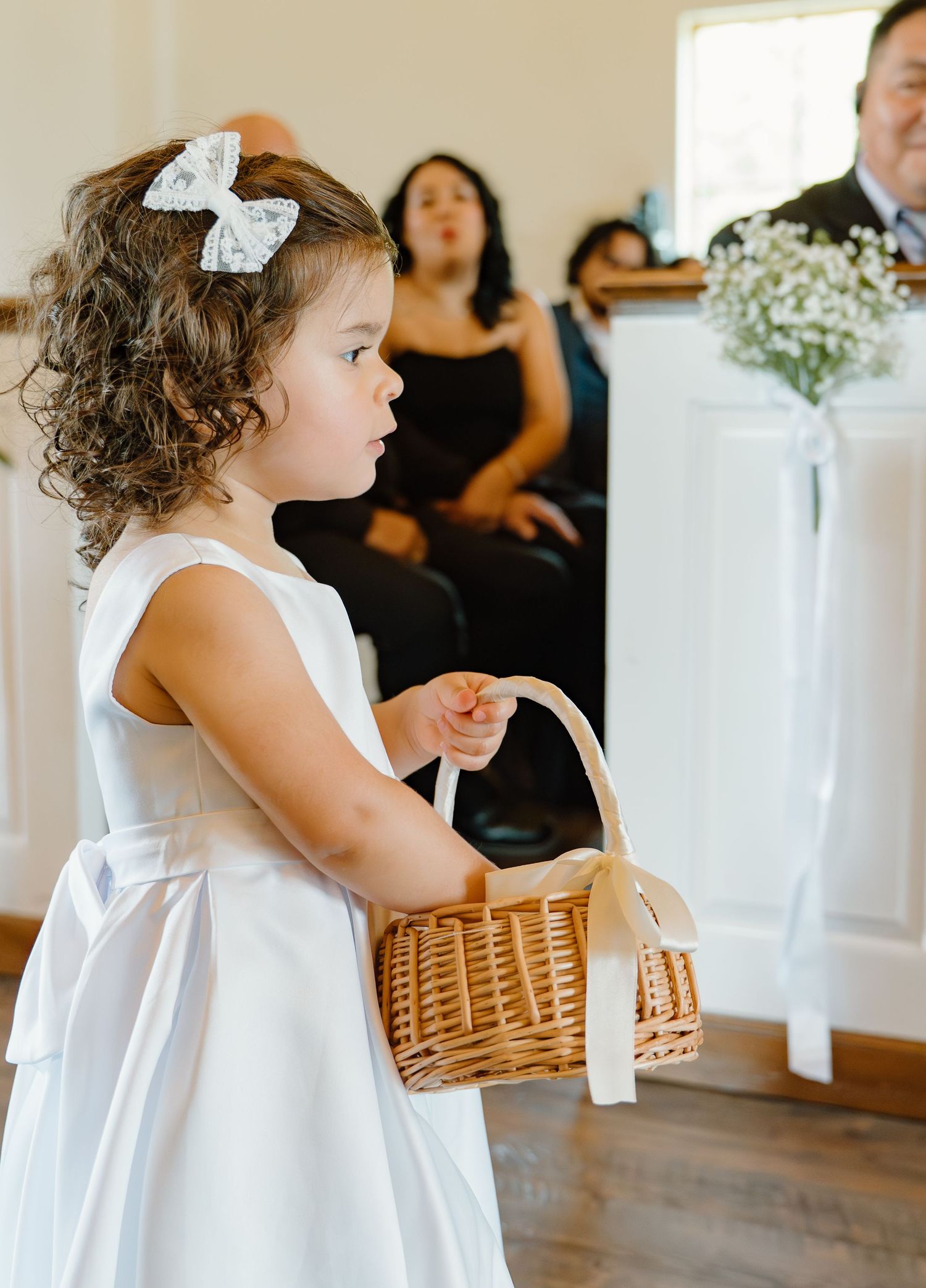 Adorable curly-haired flower girl in white dress carrying wicker basket down wedding ceremony aisle.